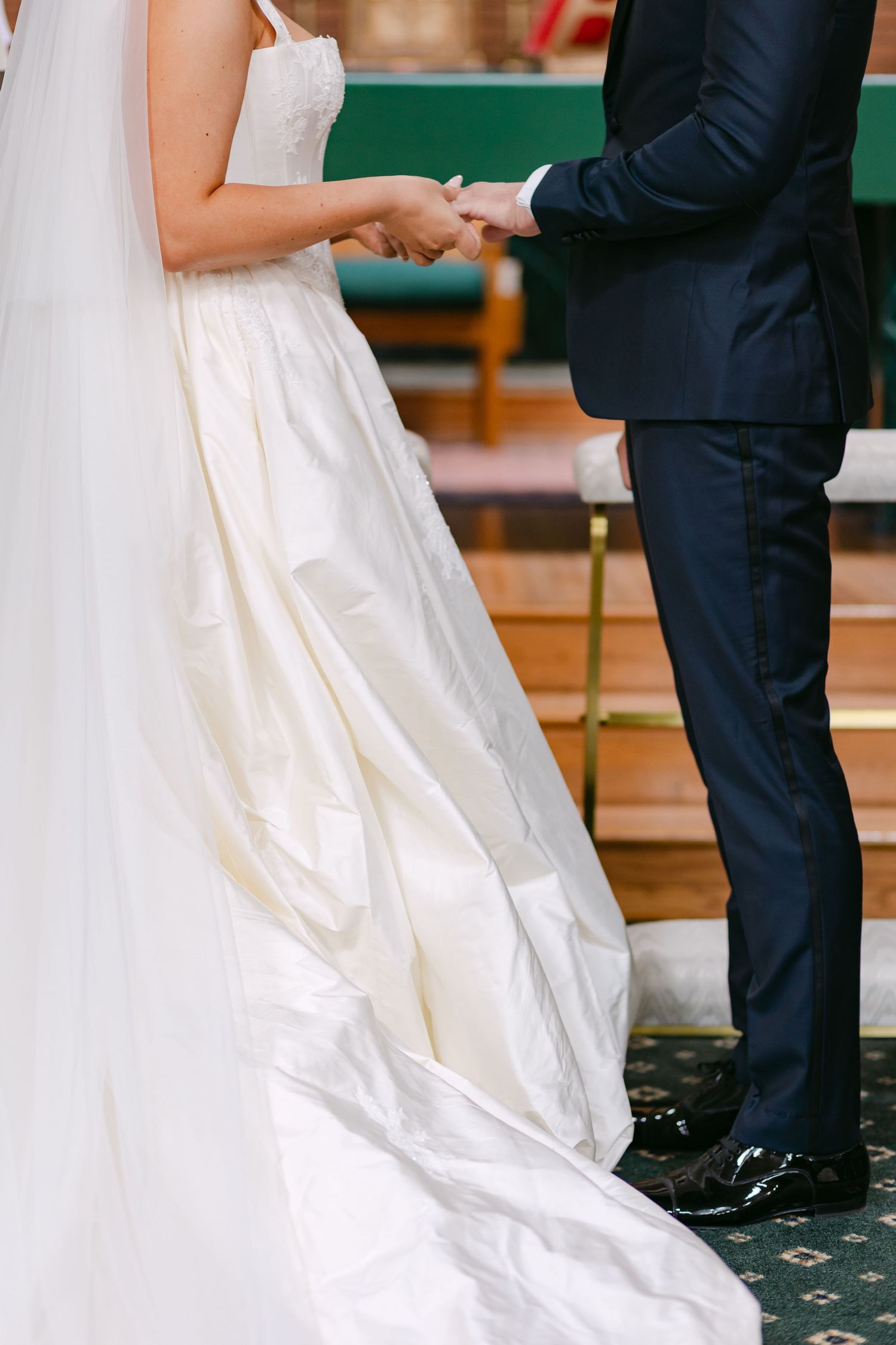 Bride and groom holding hands during wedding ceremony, bride in elegant white gown with long train in church.