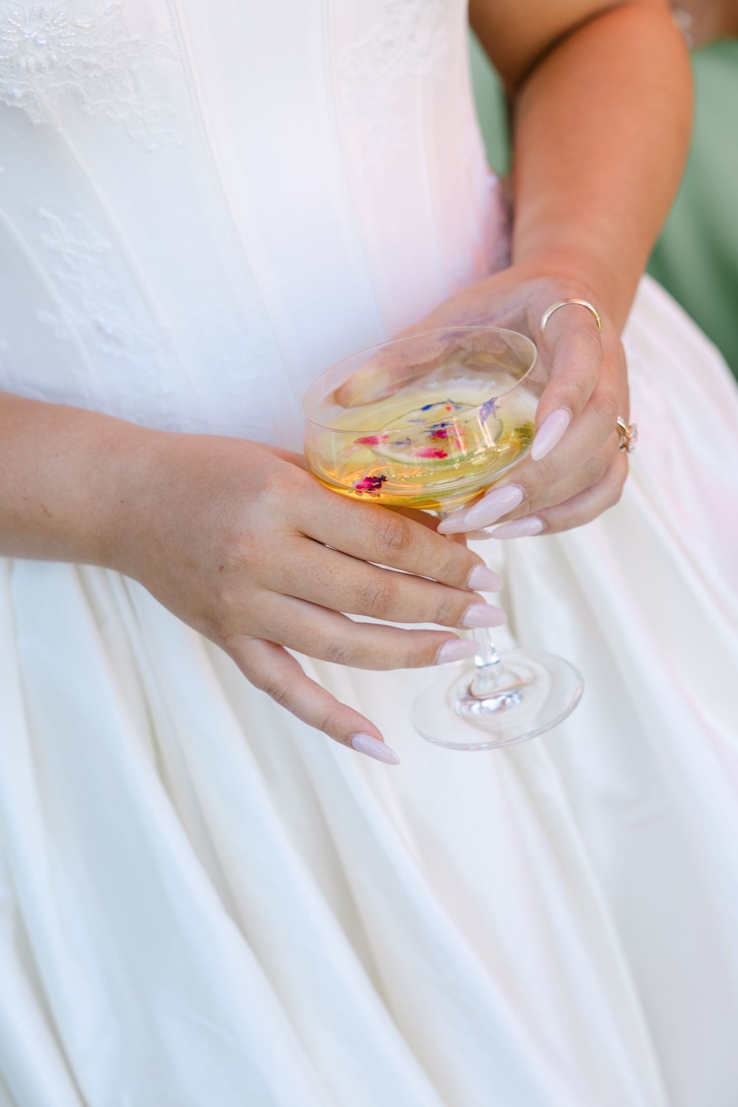 Bride in white lace gown holds a wine glass with floral garnish, showing elegant long nails and a delicate ring.