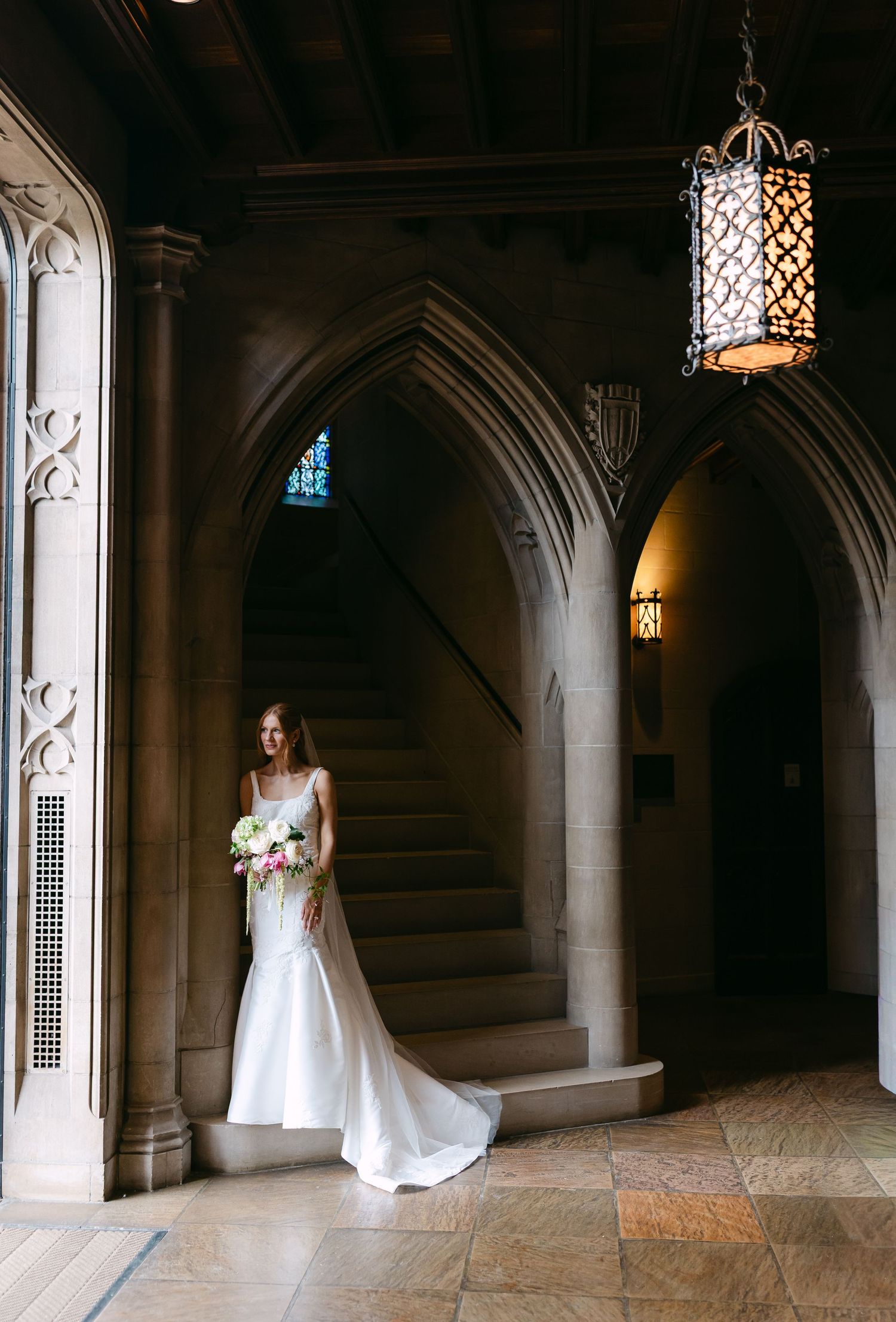 Bride in fitted white gown holds bouquet in Gothic church interior with arched stone staircase.