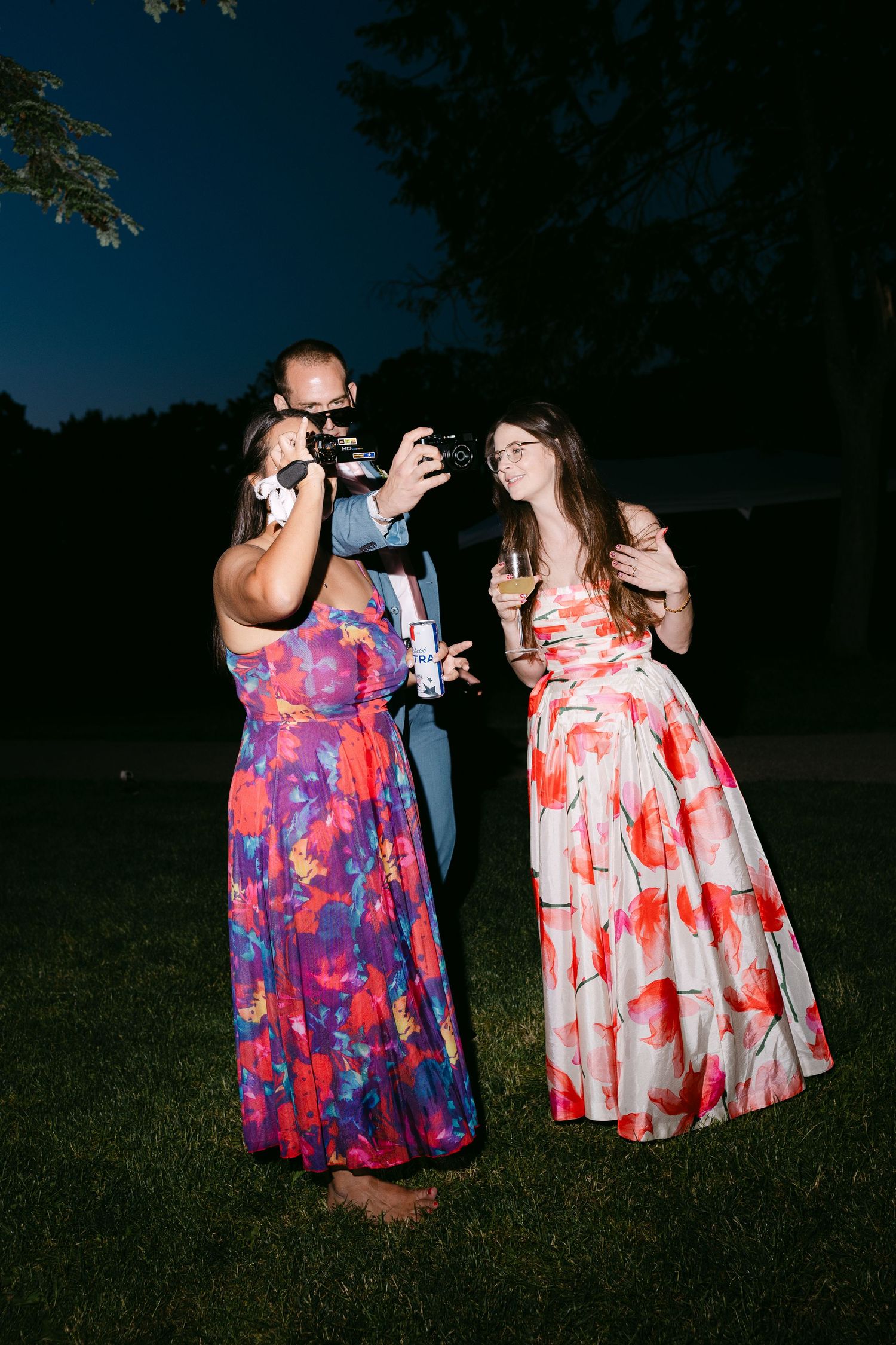 Two women in colorful floral gowns pose for a camera photo at an outdoor evening wedding reception.