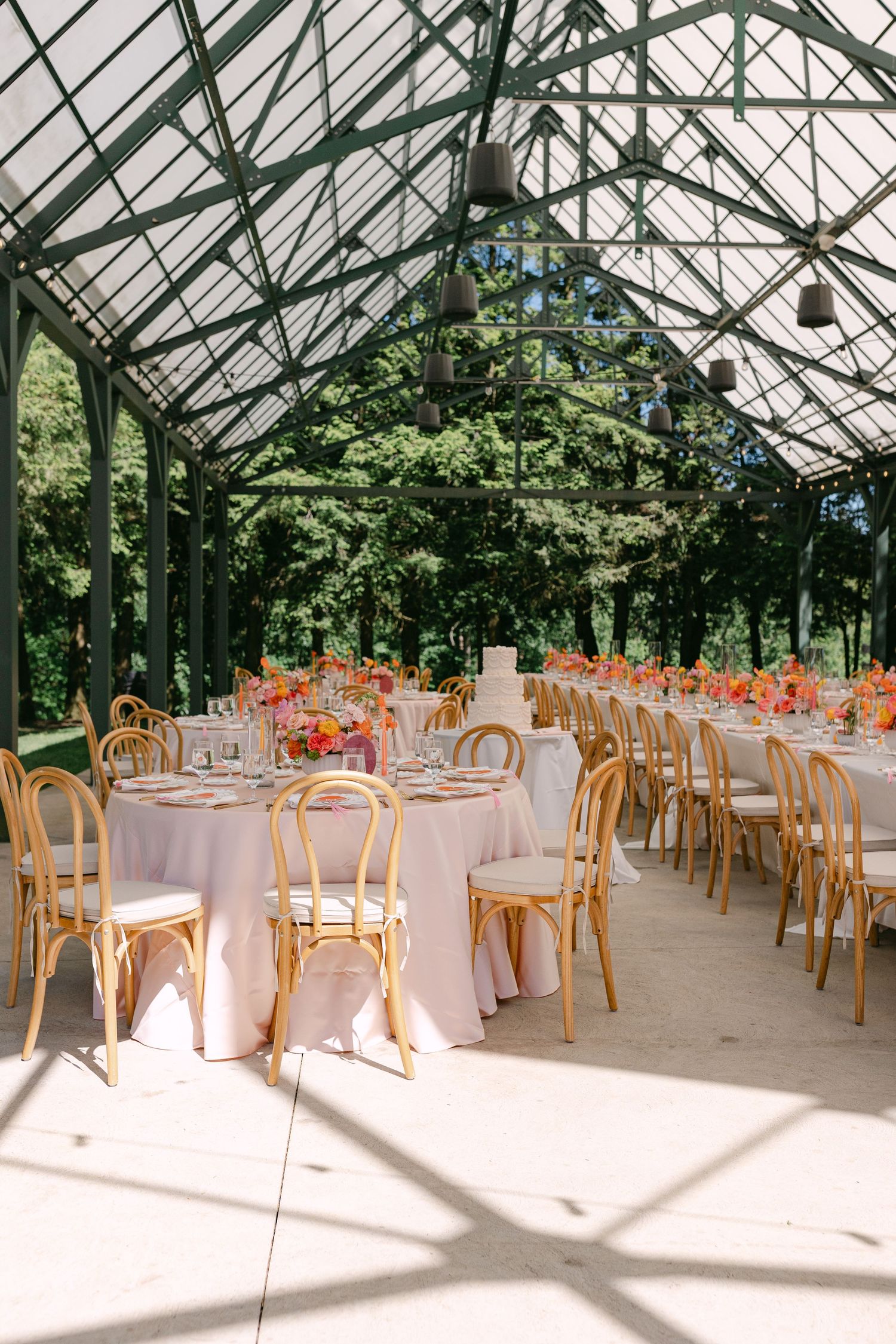 Elegant greenhouse wedding reception with bentwood chairs, pink tablecloths, and vibrant floral centerpieces under glass ceiling.