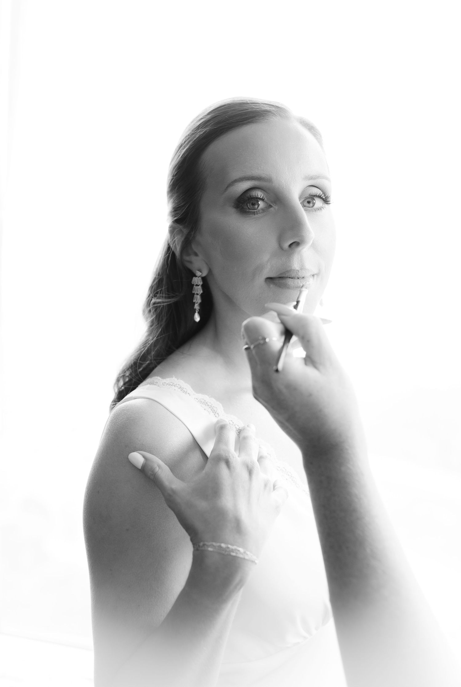 Black and white portrait of a bride having her makeup applied, wearing elegant drop earrings and a white dress.