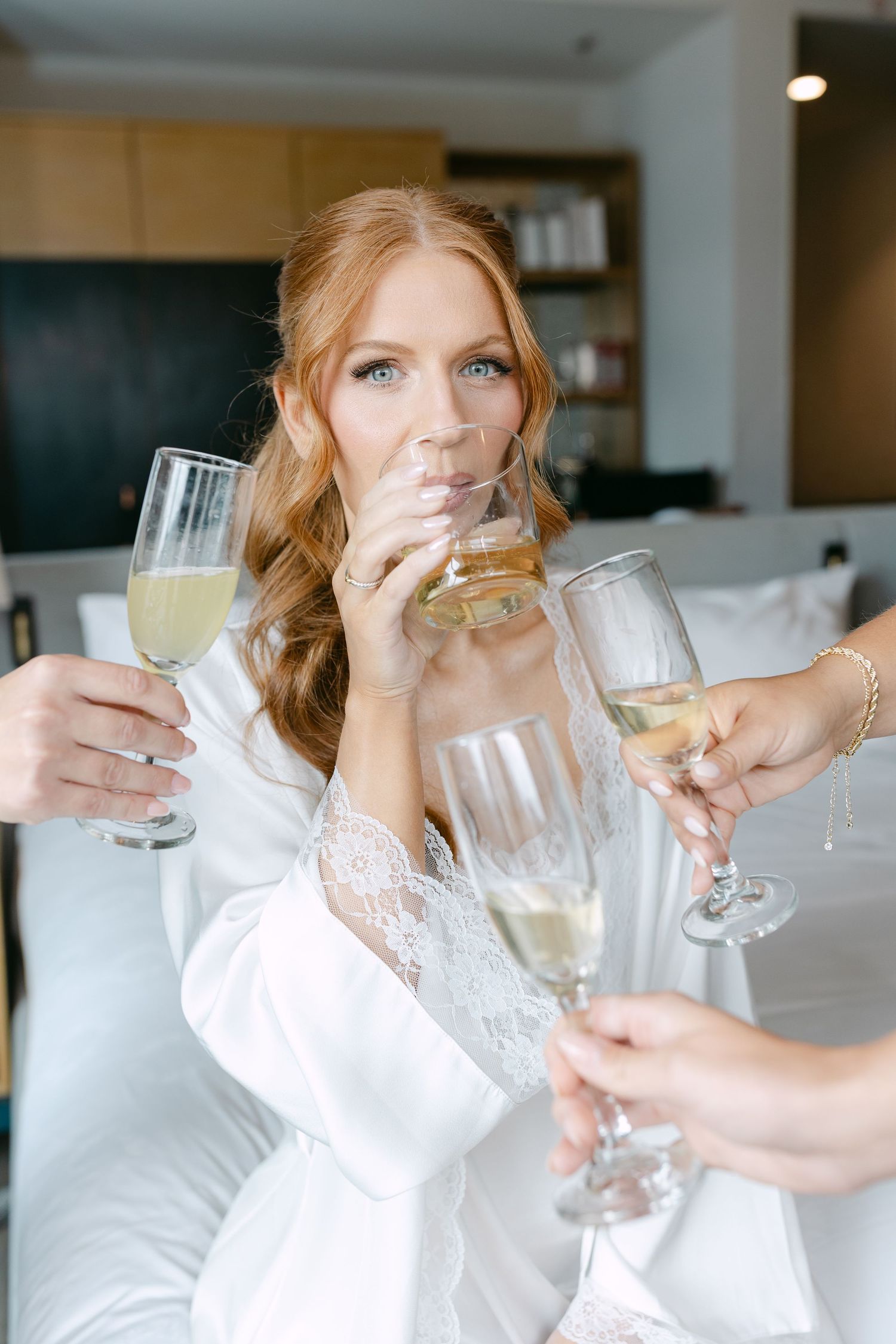 Redhead bride in white robe toasts champagne with bridesmaids during wedding morning preparations.