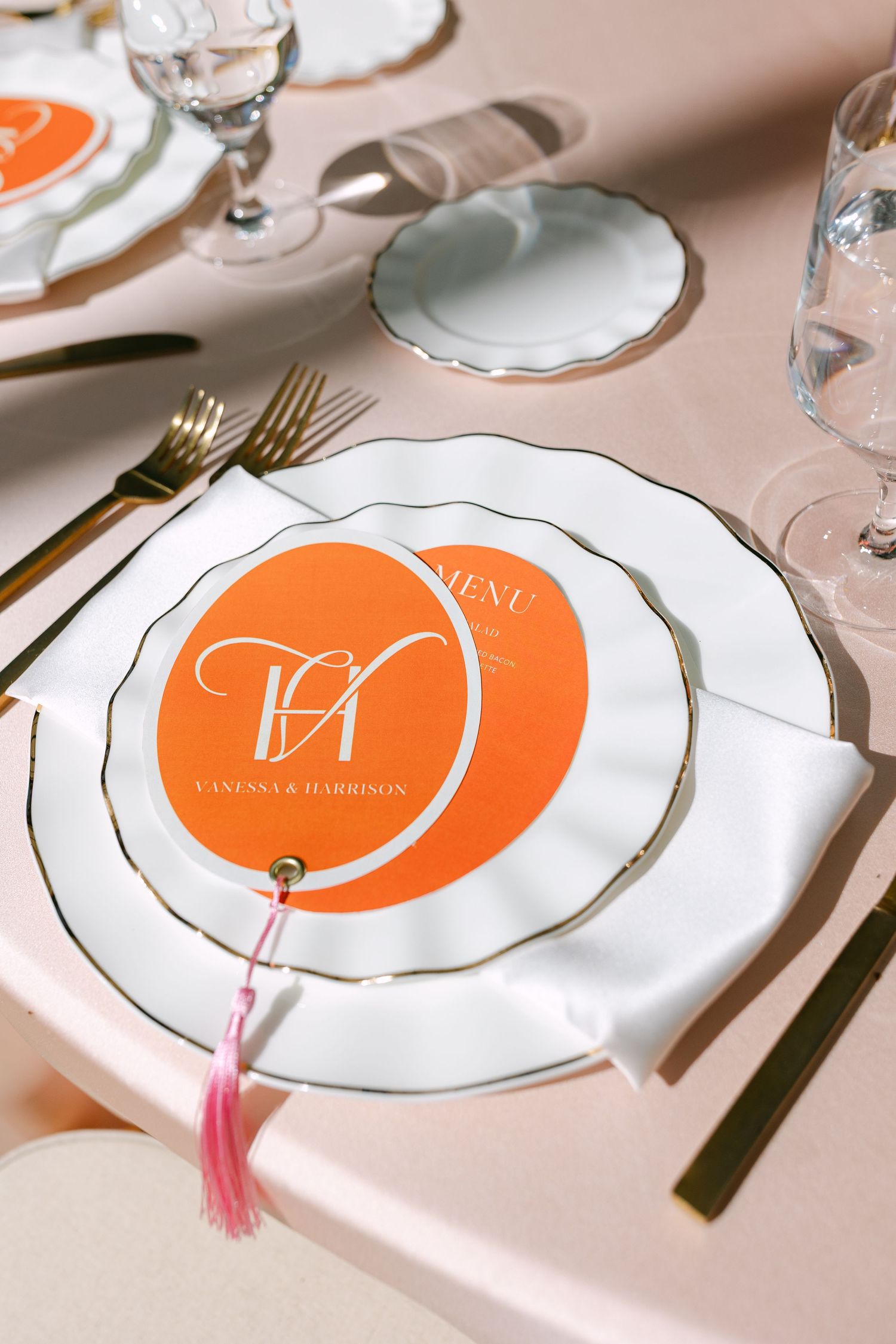 Orange circular wedding menu card with monogram on white scalloped plate with gold cutlery place setting.