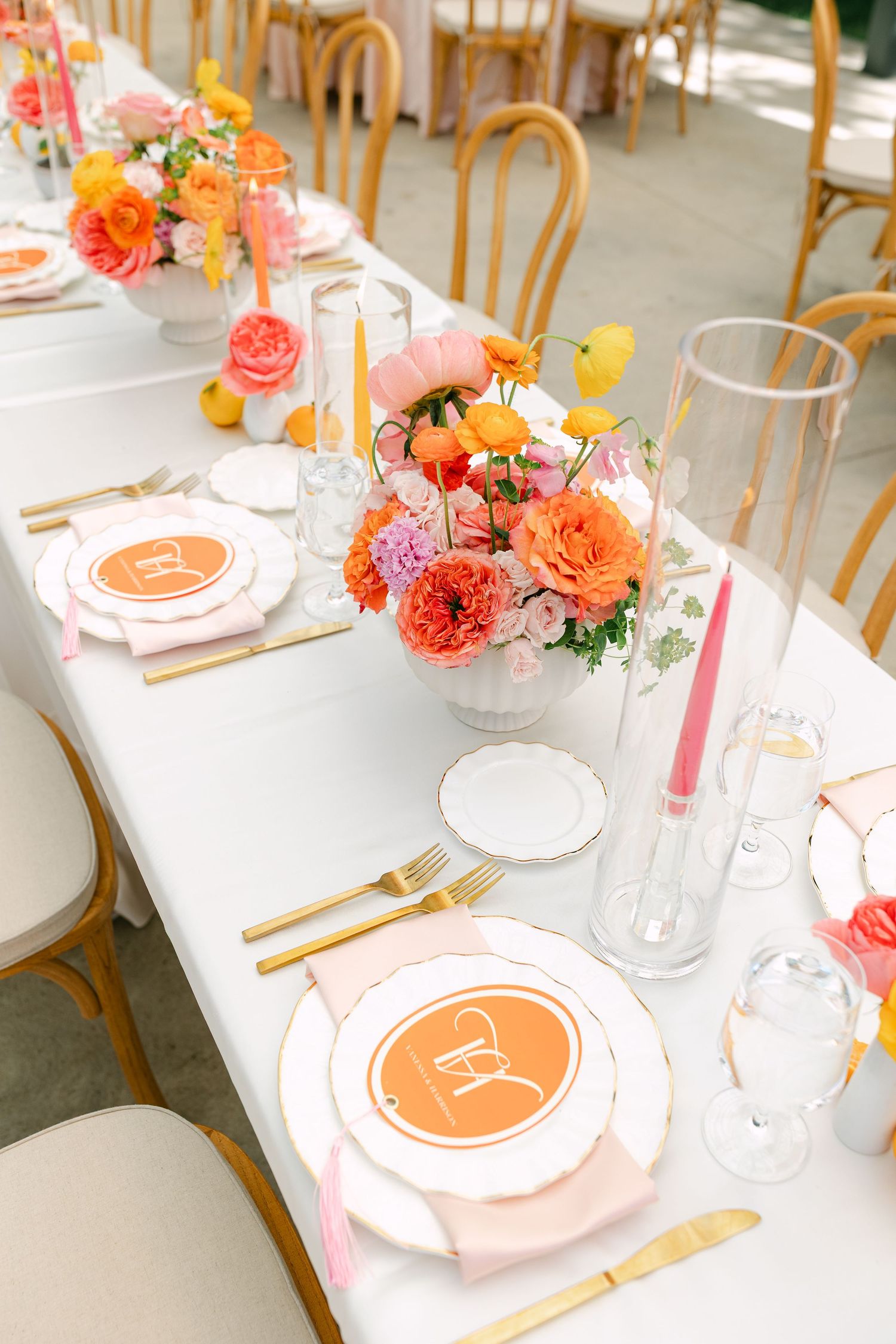 Colorful wedding reception table setting with orange floral centerpieces, pink napkins, gold flatware, and personalized place settings.