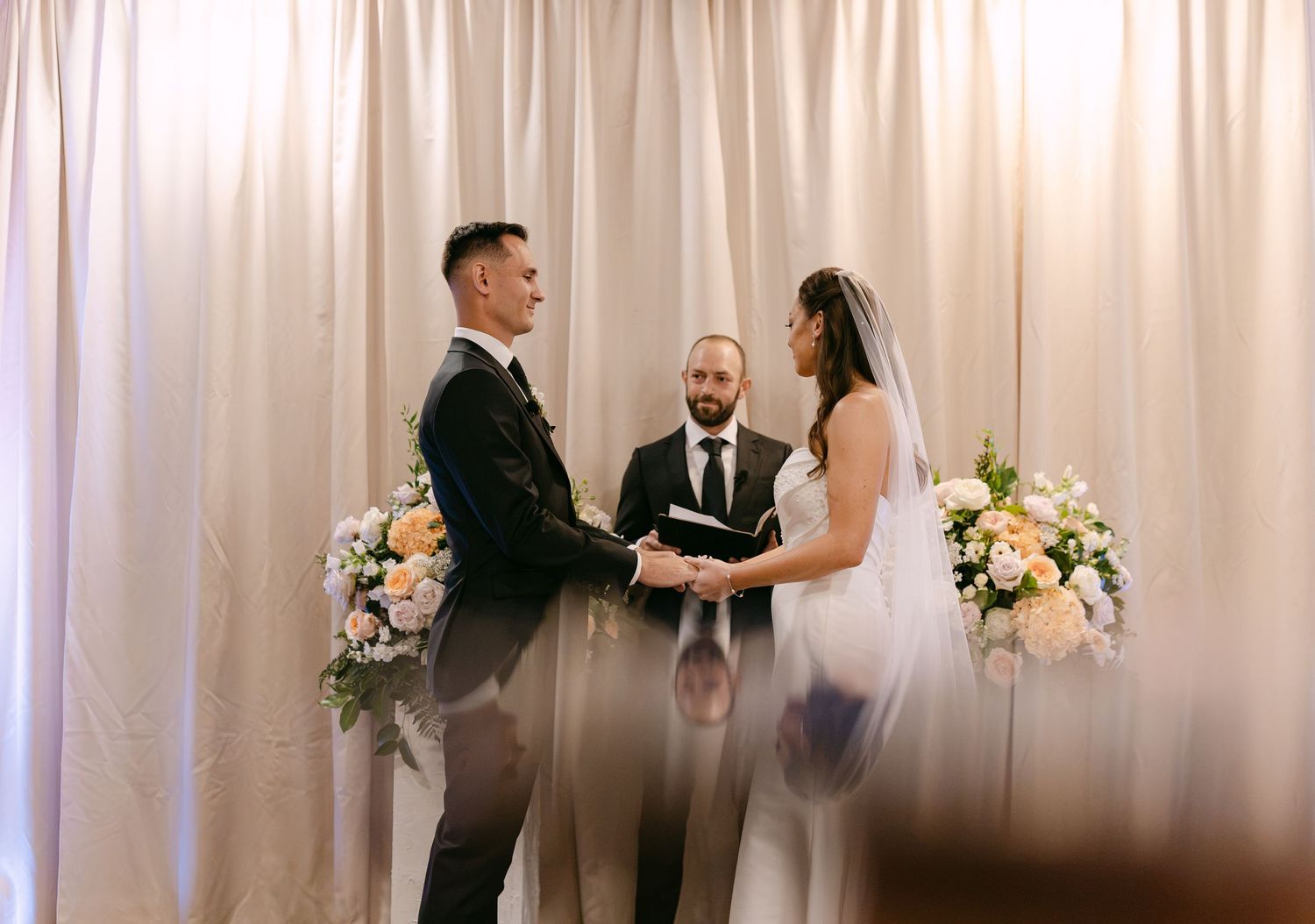 Groom and bride hold hands at intimate ceremony altar with white draping and pastel floral arrangements.