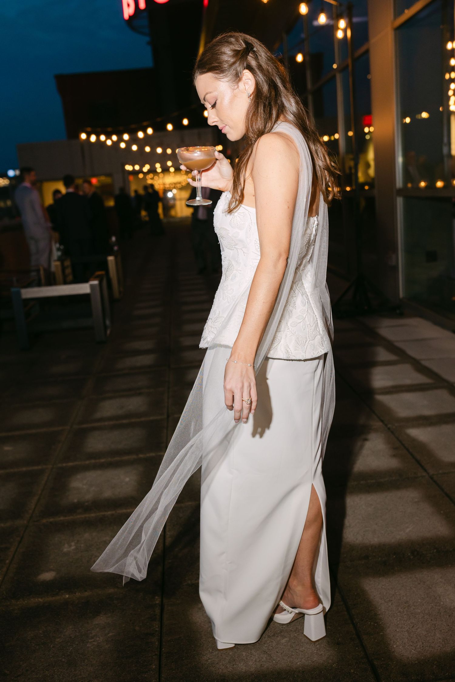 Elegant bride in white peplum top and skirt holding a cocktail glass on a rooftop venue at night.