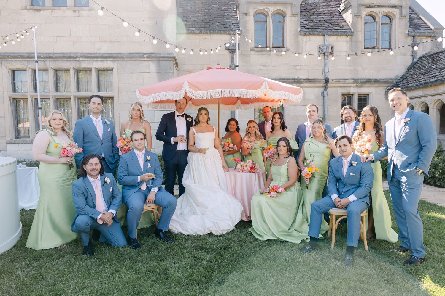Wedding party group photo with bride and groom, bridesmaids in green, and groomsmen in blue outside stone manor.