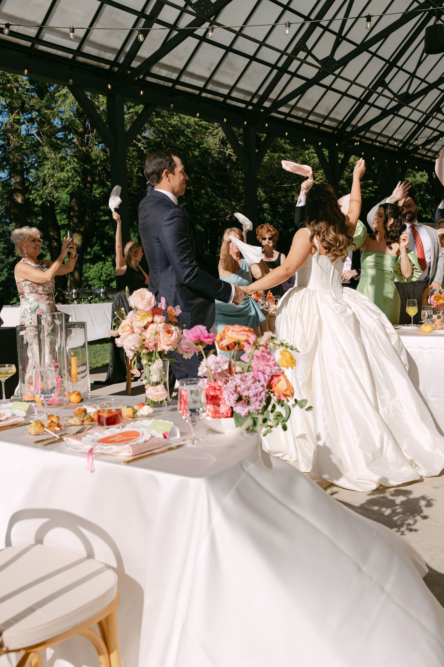 Bride and groom celebrate with guests waving napkins at a floral-decorated outdoor wedding reception table.