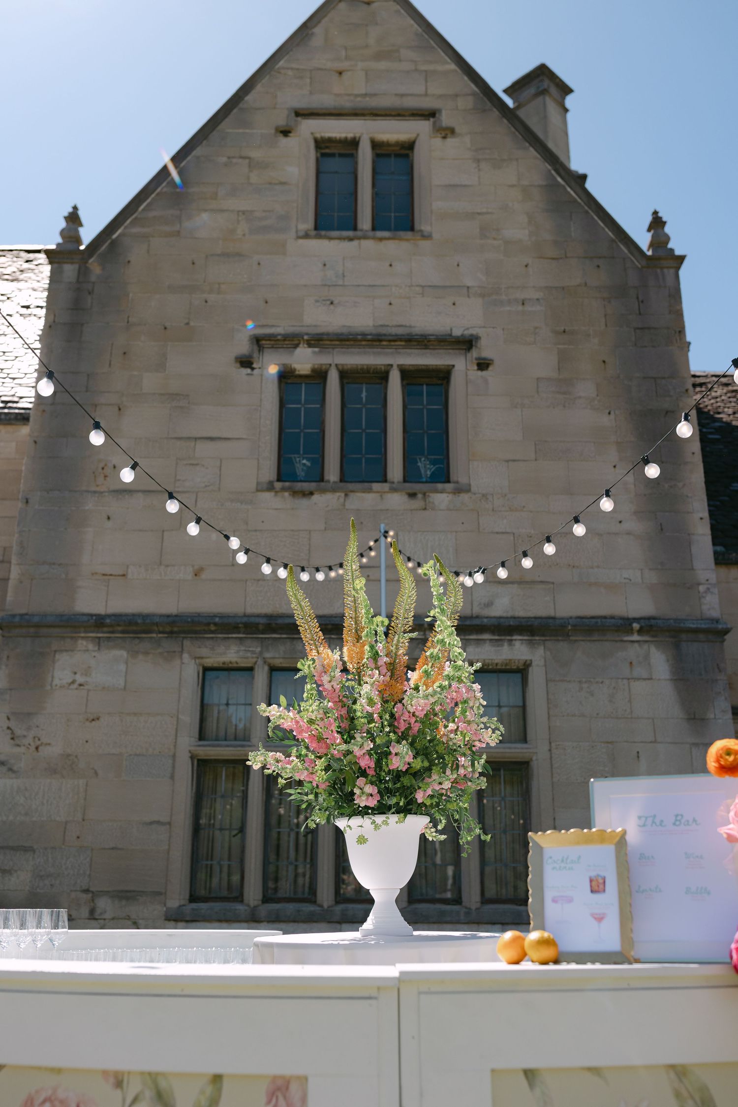 Elegant floral arrangement in white urn displayed on outdoor bar at stone manor wedding venue with festoon lights.