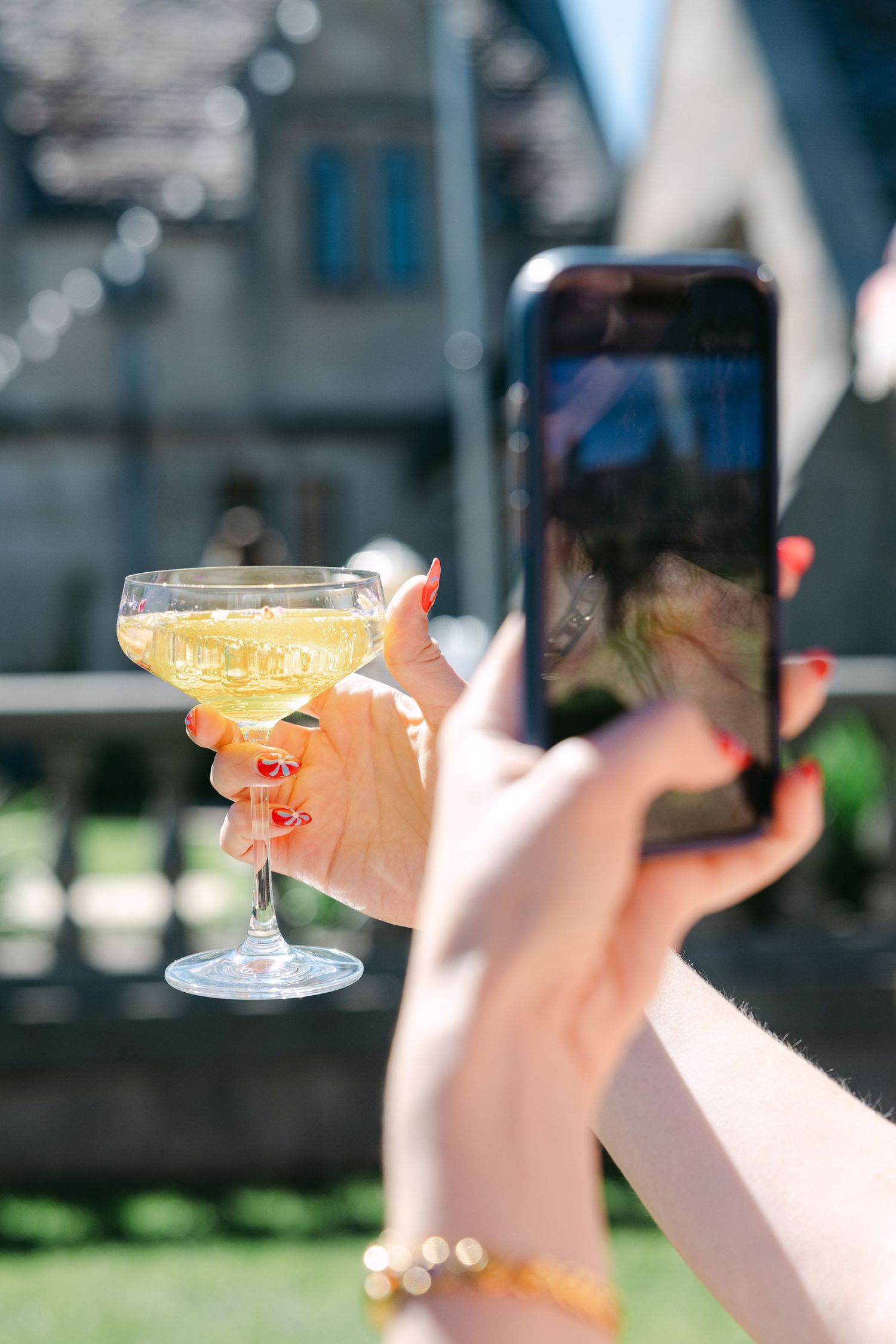 Woman with red nails photographing a champagne coupe cocktail glass outdoors with a smartphone.