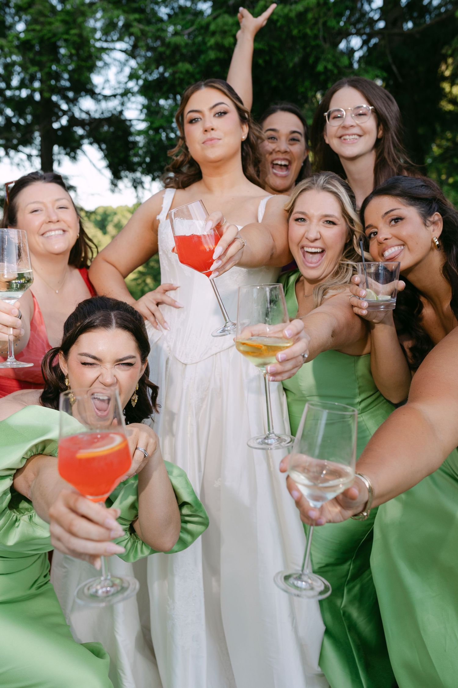 Bride in white gown celebrating with bridesmaids in green satin dresses holding colorful cocktails outdoors.