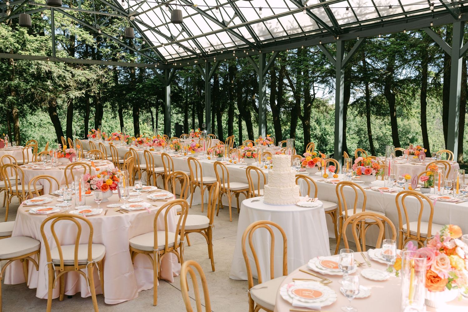 Elegant wedding reception in a glass greenhouse with round tables, gold chairs, and vibrant pink and orange floral centerpieces.