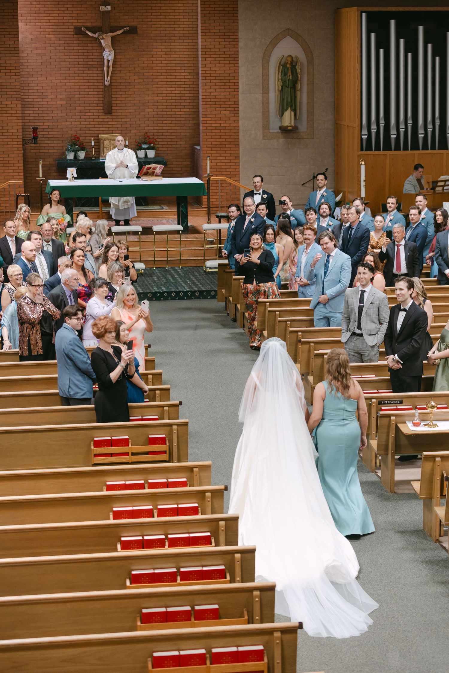 Bride walks down the aisle in a Catholic church wedding ceremony, guests standing in wooden pews.