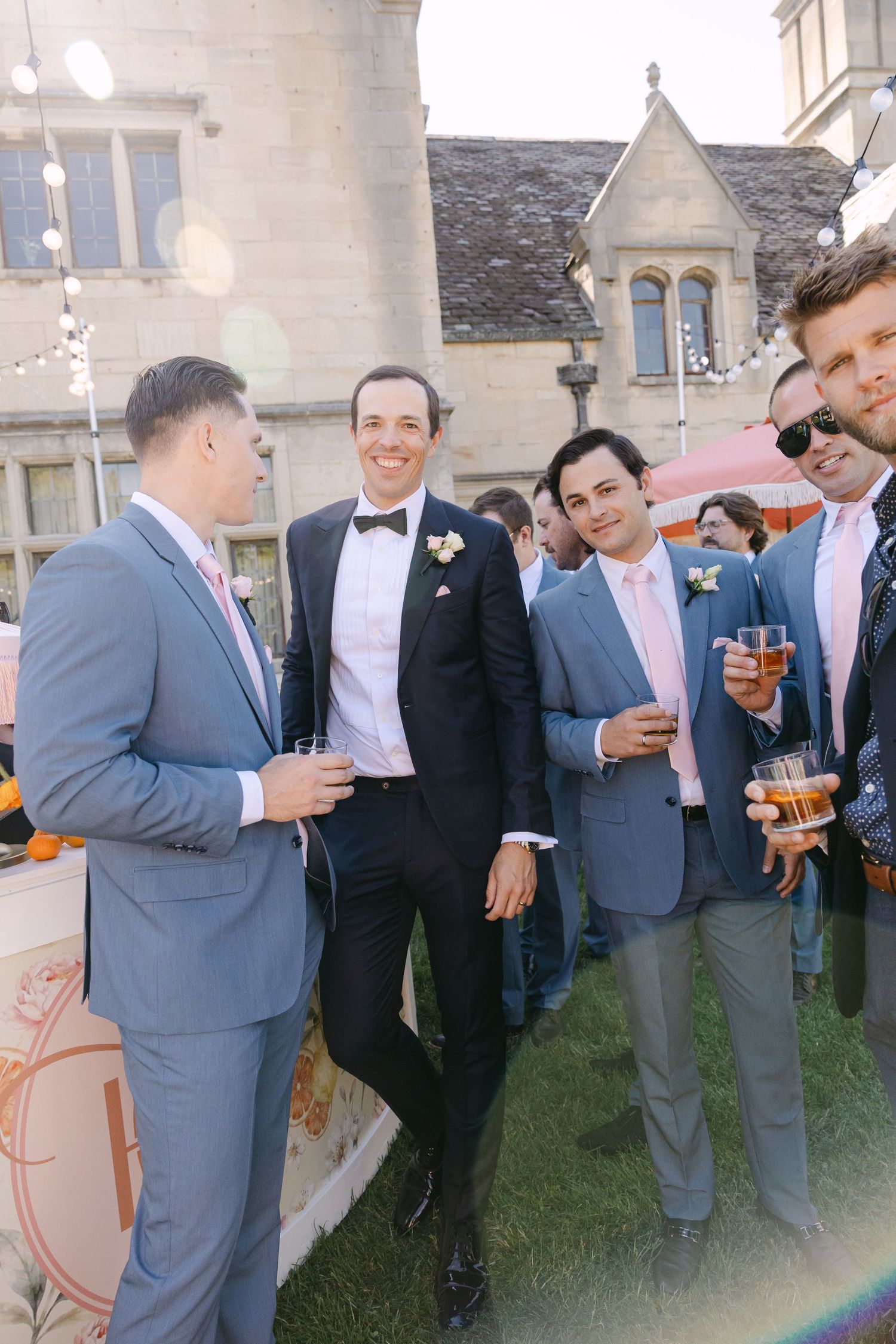 Groom in black tuxedo socializing with groomsmen in grey suits at an outdoor wedding cocktail hour.