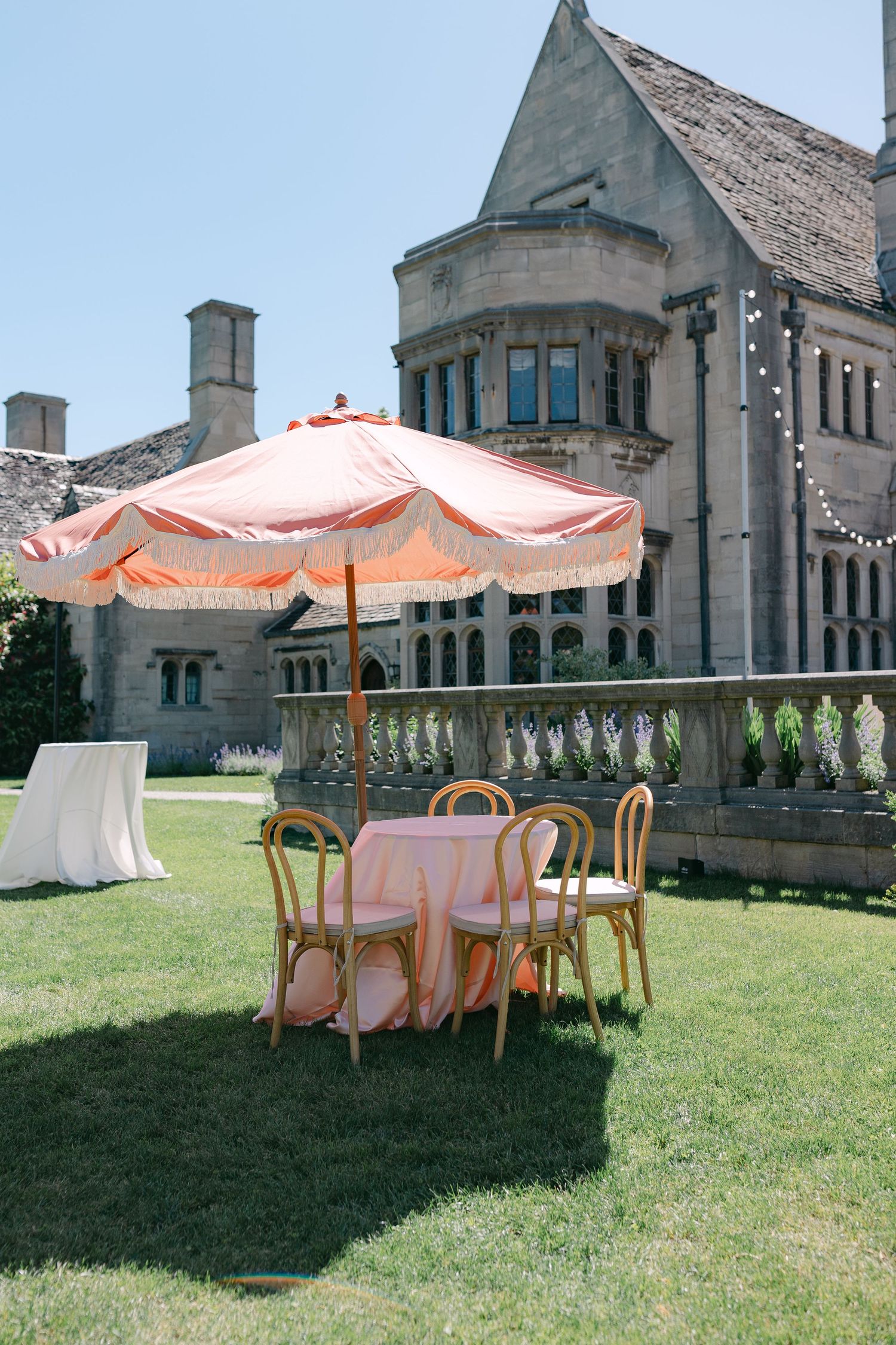 Elegant outdoor wedding reception setup with peach umbrella and chairs on lawn of stone mansion.
