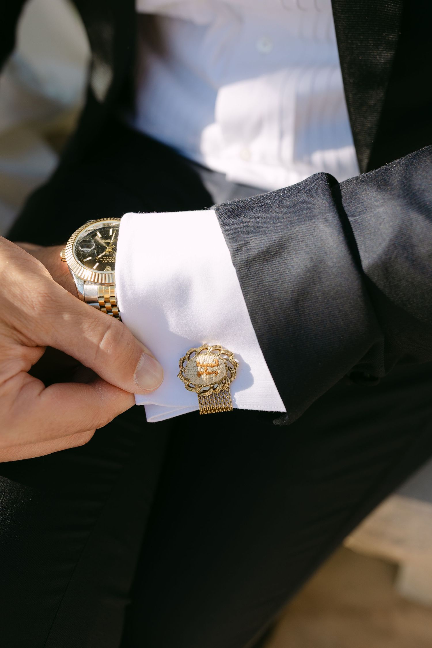 Groom adjusting gold lion cufflink on white dress shirt with gold Rolex watch in black tuxedo.