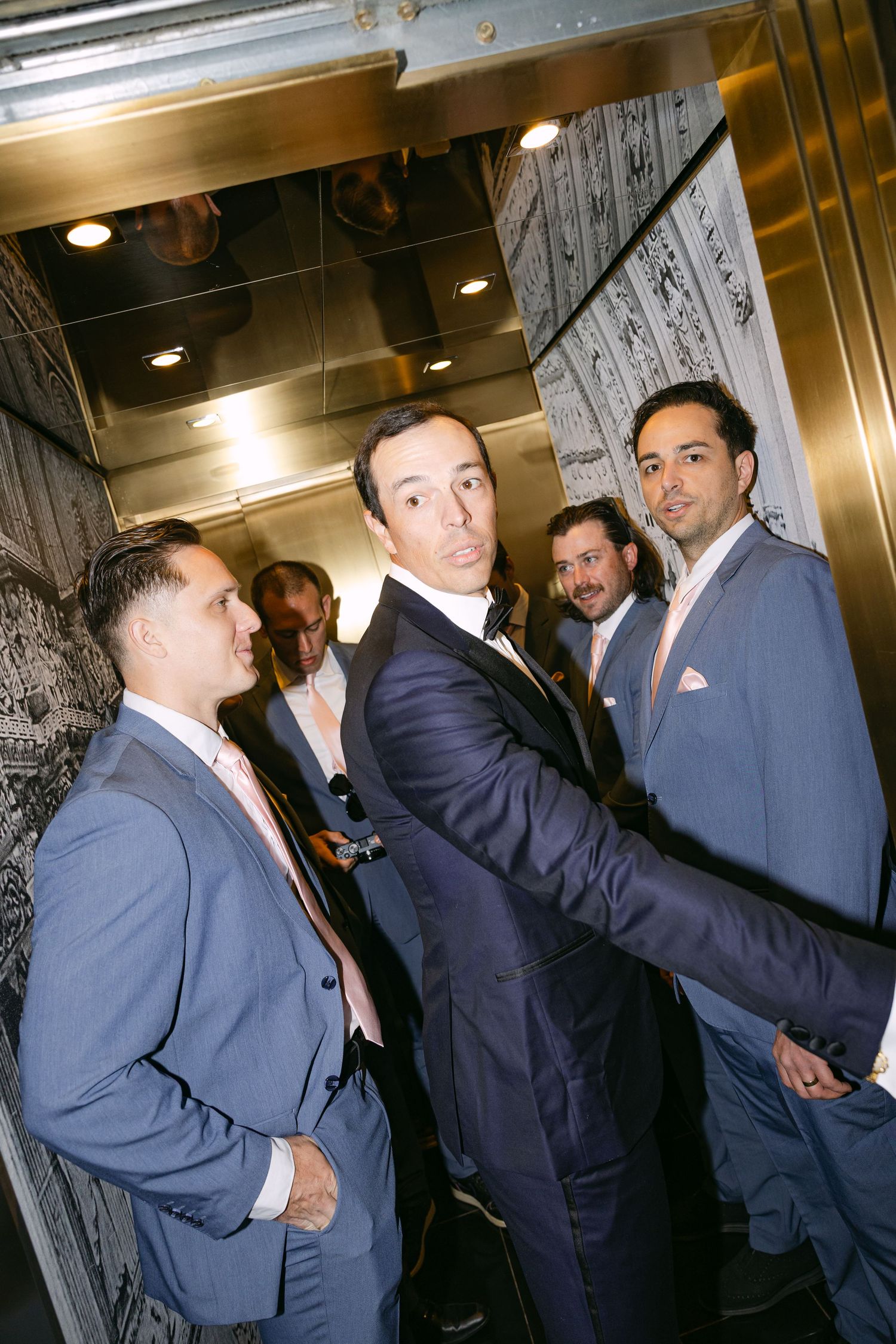 Groomsmen in navy and blue suits posing together inside a mirrored elevator at a wedding celebration.