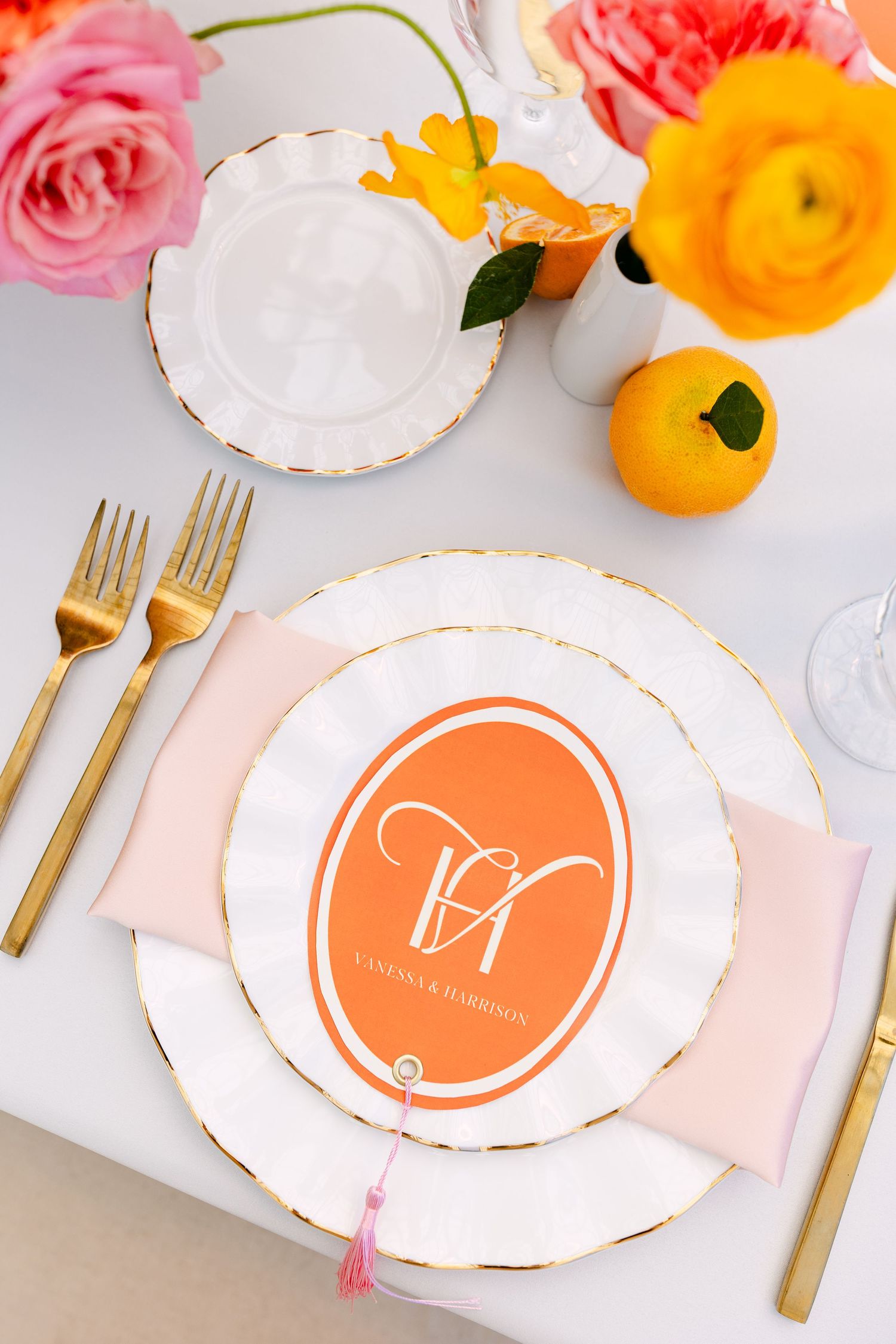 Elegant wedding place setting with orange monogram card, gold cutlery, blush napkin, and fresh flowers on a white tablecloth.