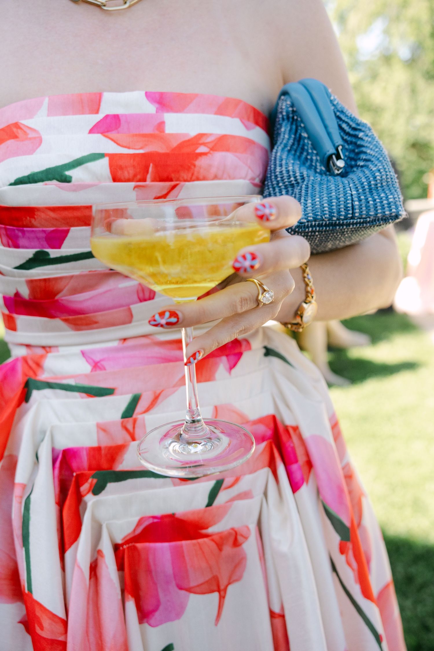 Guest in floral strapless dress holding yellow cocktail and blue beaded clutch at outdoor wedding.