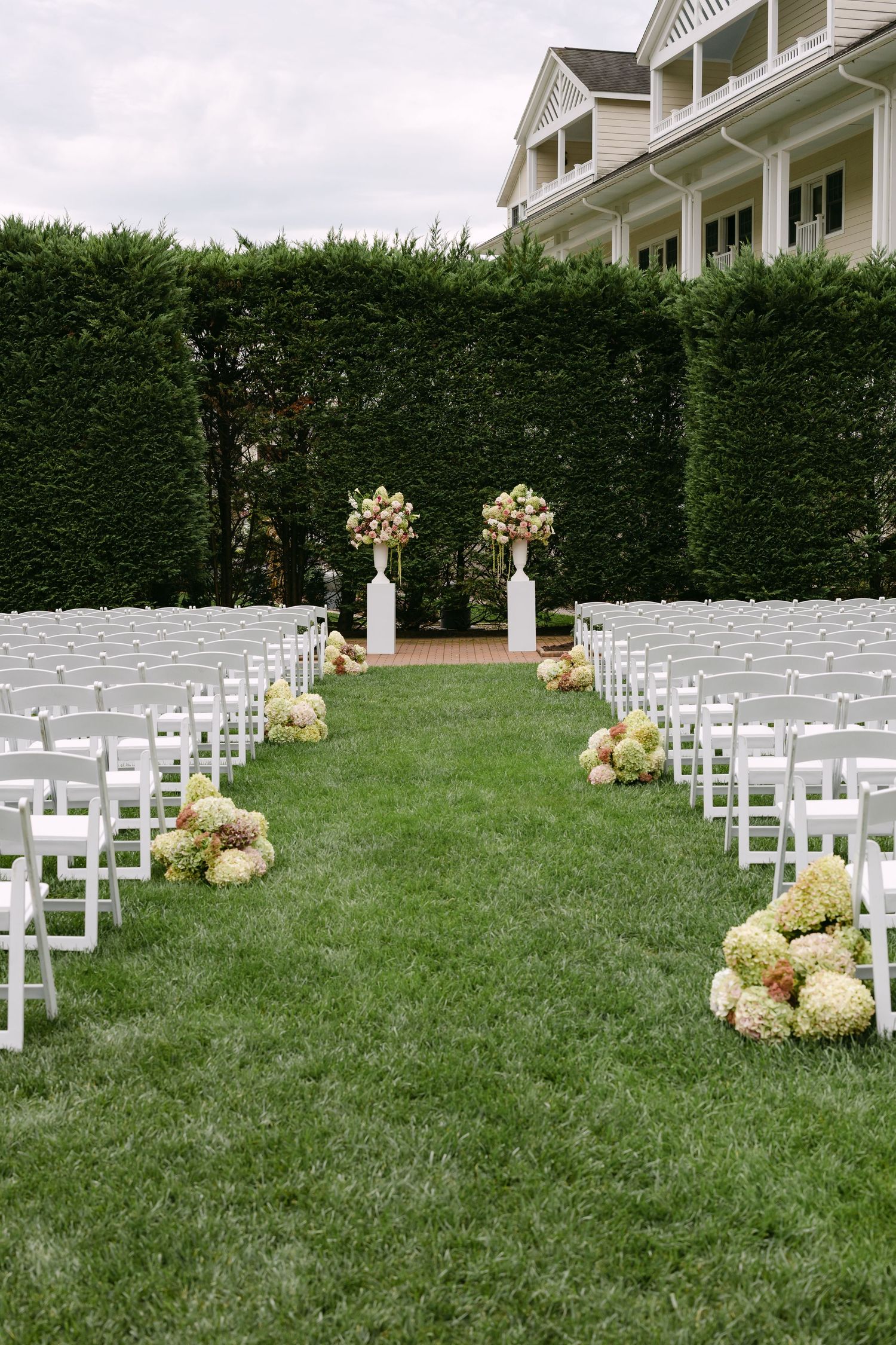 Elegant outdoor wedding ceremony setup with white chairs, hydrangea aisle flowers, tall floral arrangements, and manicured hedges.