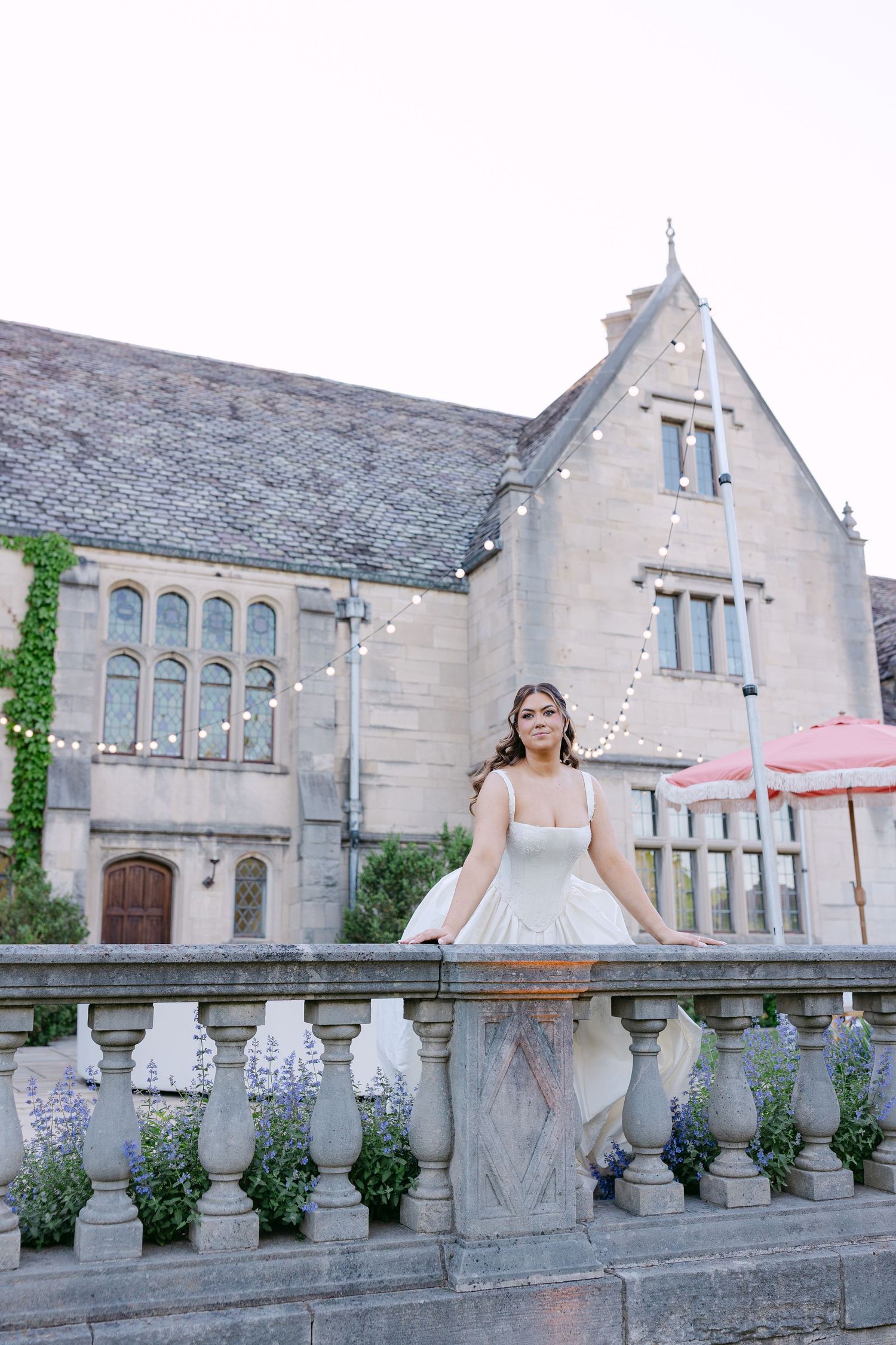 Elegant bride in white corset gown leans on stone balustrade outside ivy-covered historic manor with string lights.