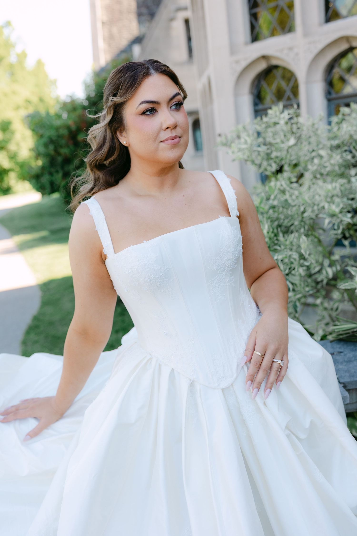 Bride in a white corset ballgown poses outdoors near a stone building with lush greenery.