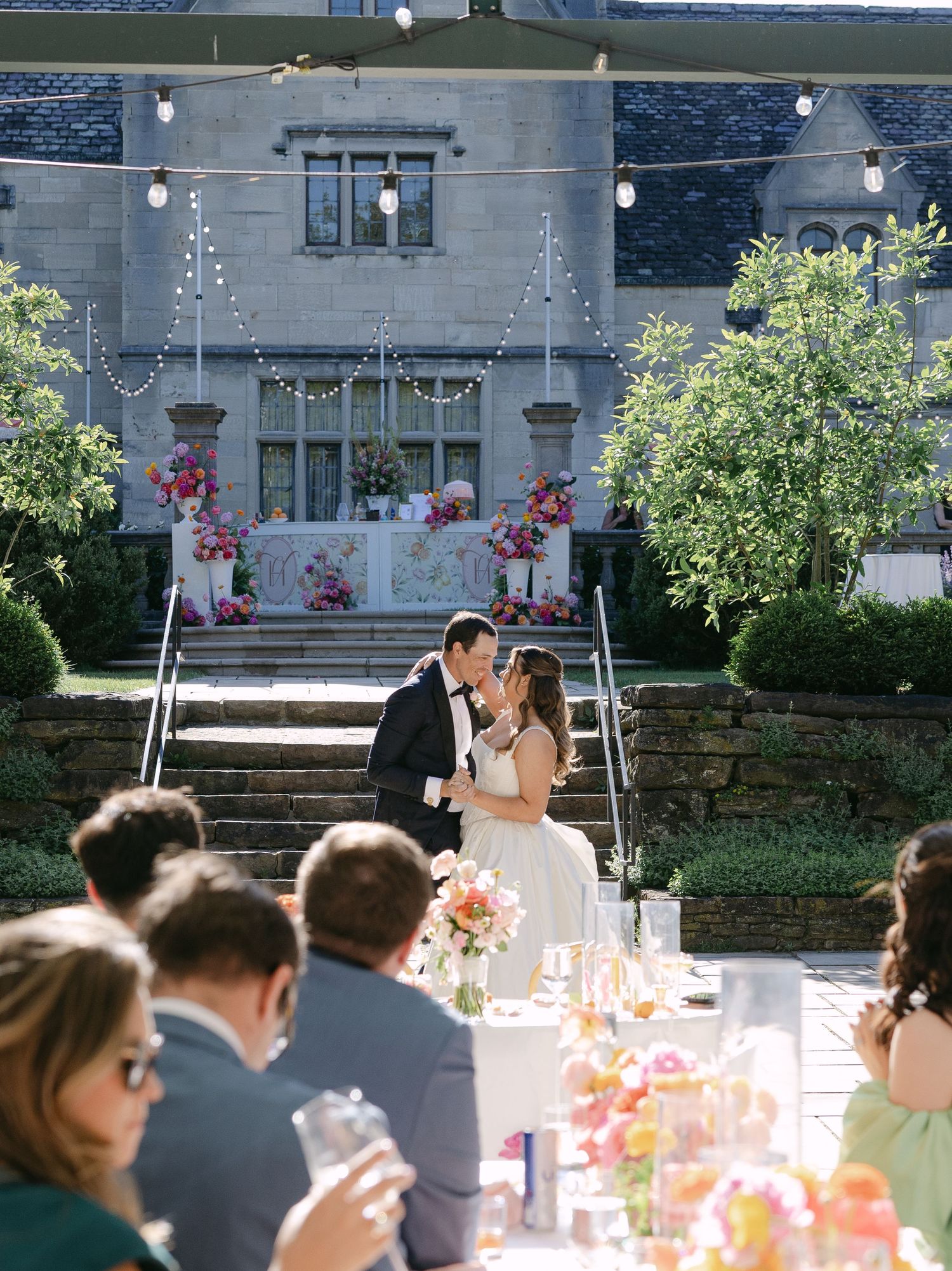 Bride and groom share a kiss on stone steps of an estate during an outdoor wedding reception.