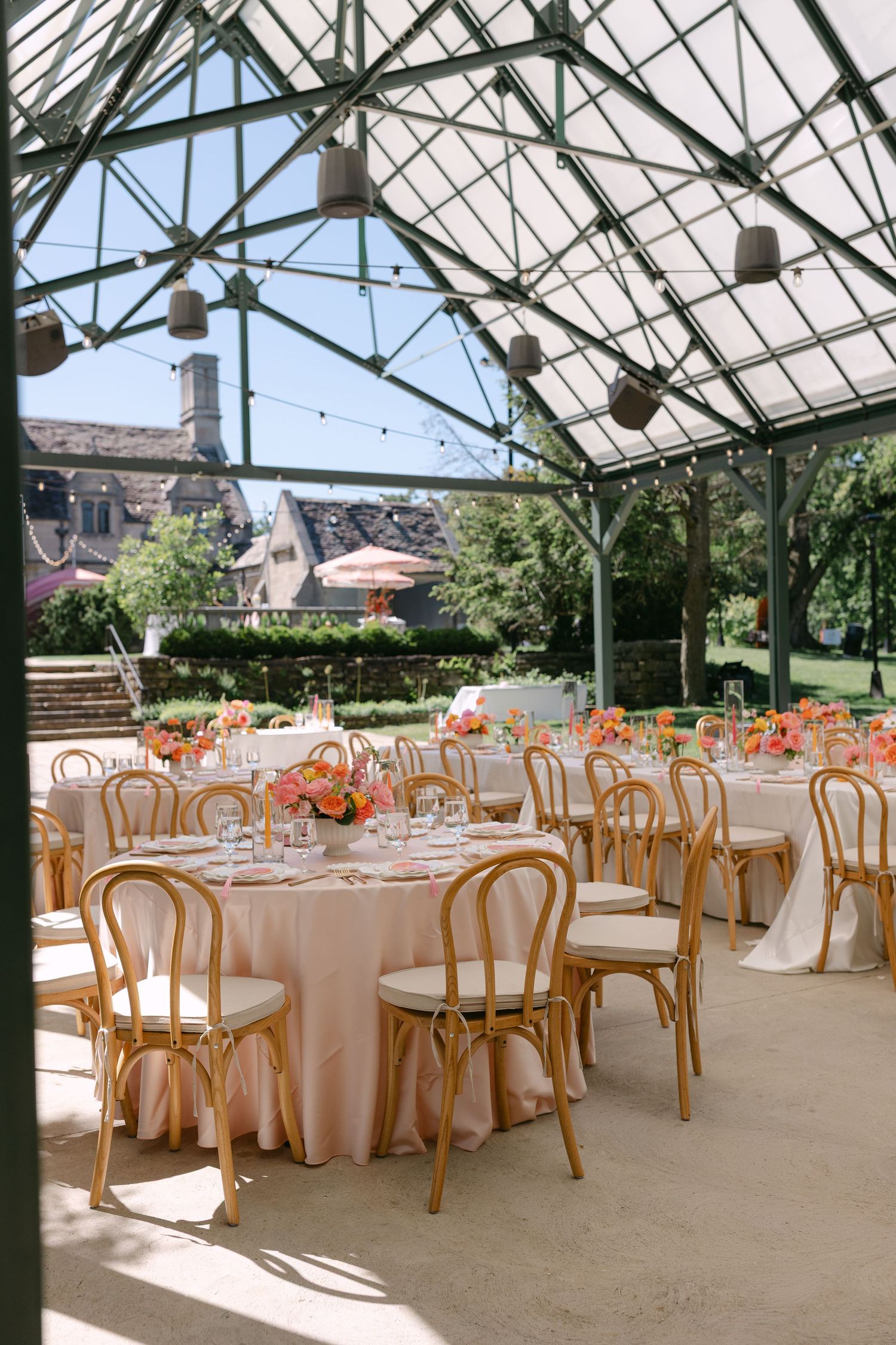 Sunlit greenhouse wedding reception with gold bentwood chairs, blush tablecloths, and bright floral centerpieces.