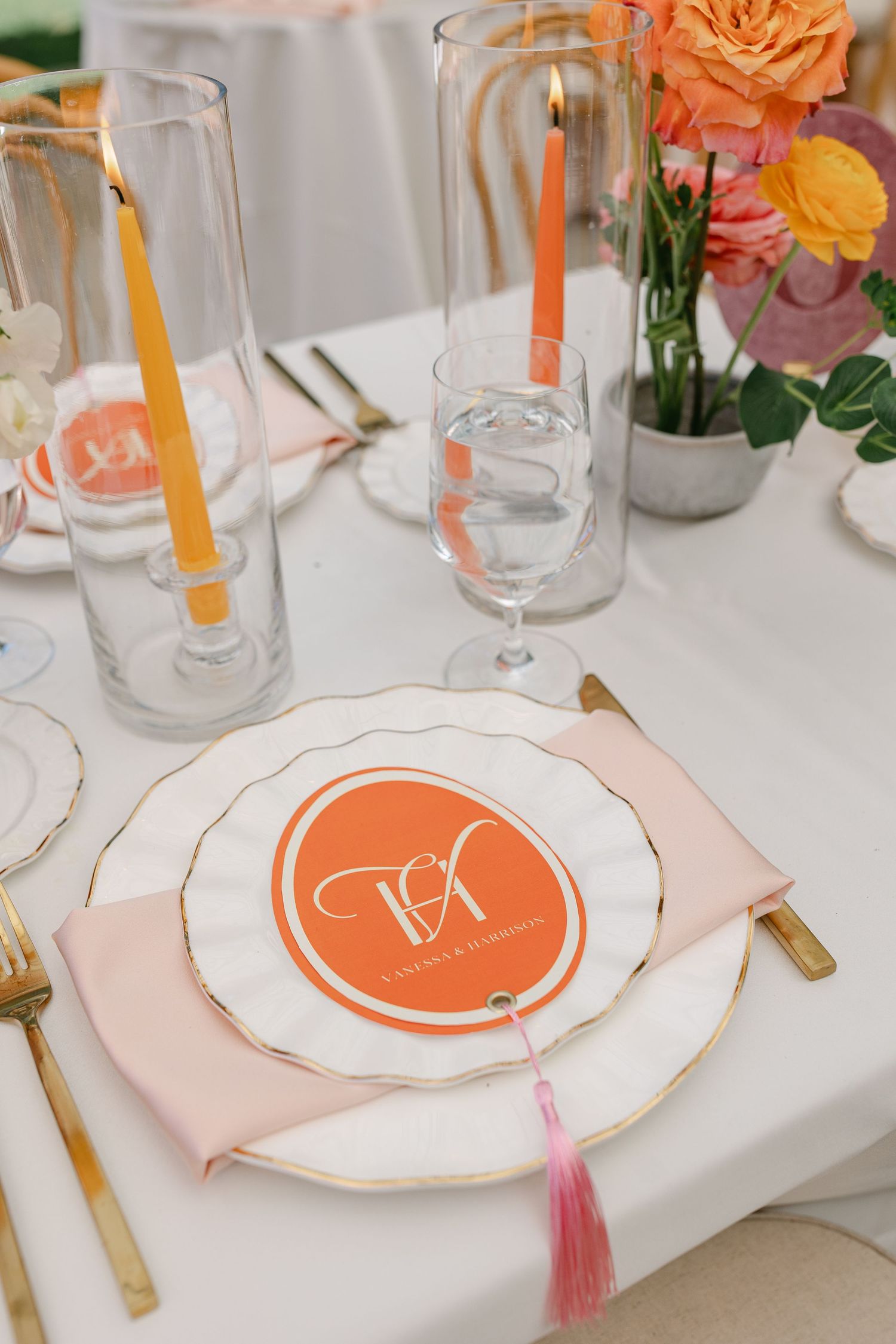 Elegant wedding place setting with orange monogram plate, gold cutlery, pink napkin, and colorful candles on white tablecloth.