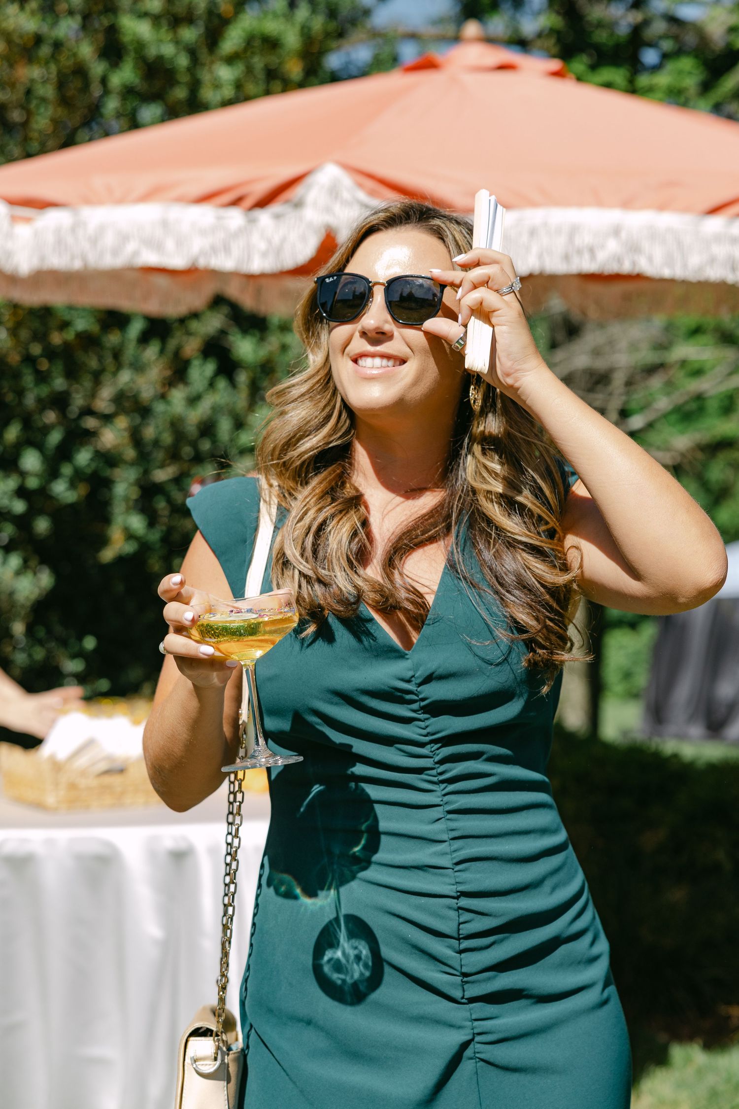 Smiling woman in green dress holding cocktail and phone under orange umbrella at outdoor wedding.