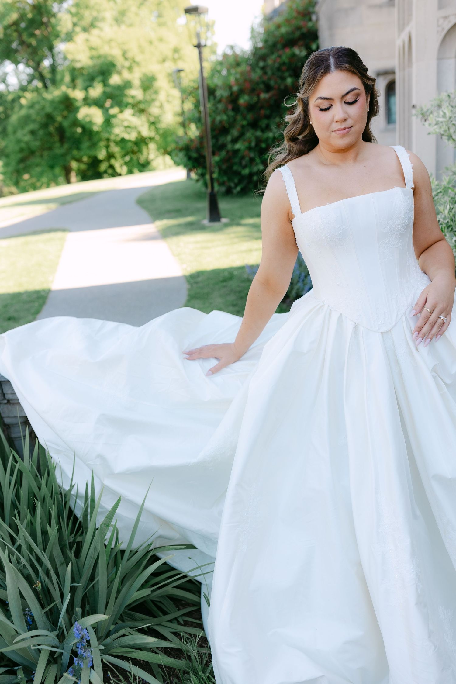 Bride in a white ball gown with square neckline and wide straps poses outdoors, flowing skirt caught in a gentle breeze.