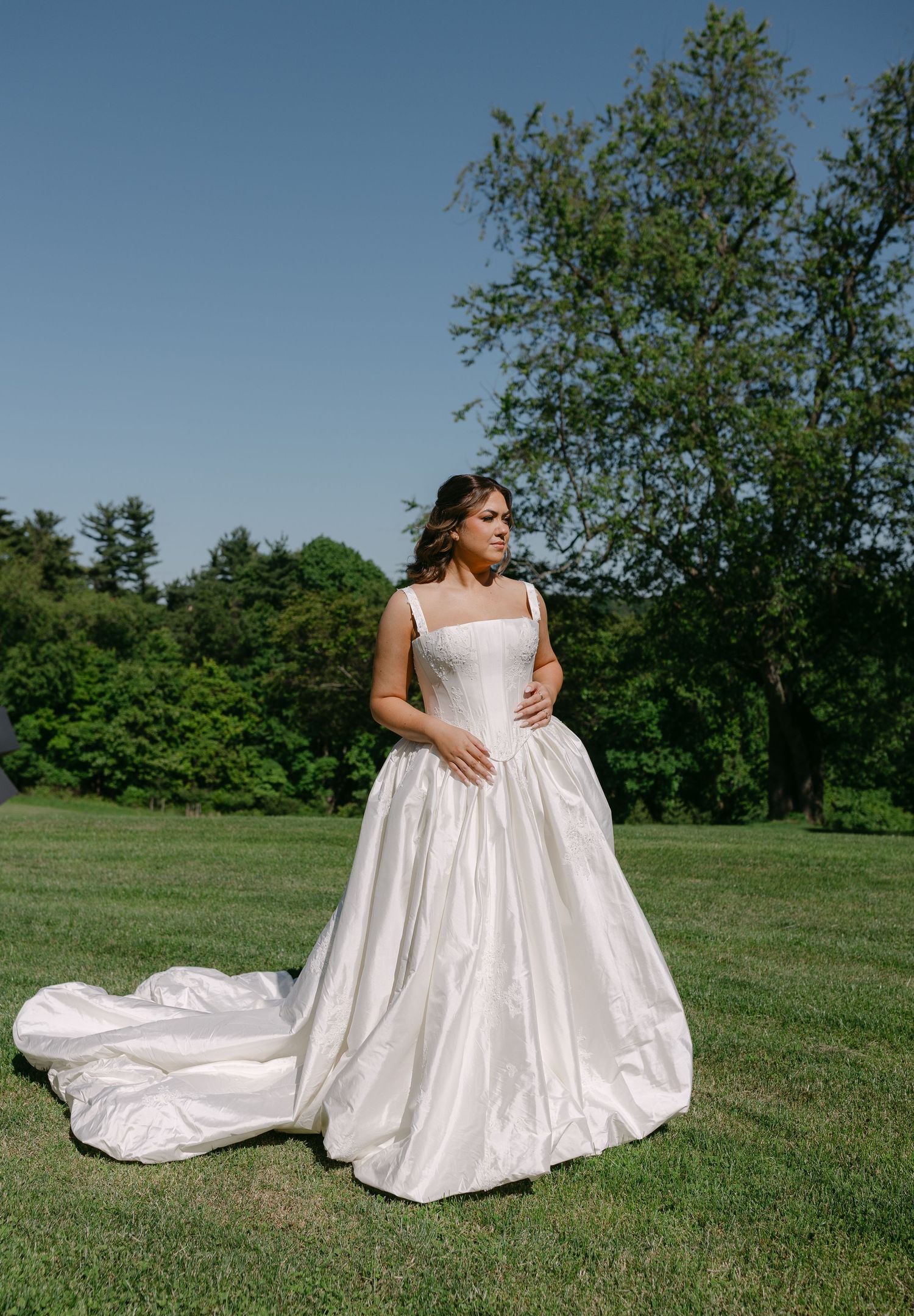 Bride in a white ballgown with long train standing on green lawn with trees in background.