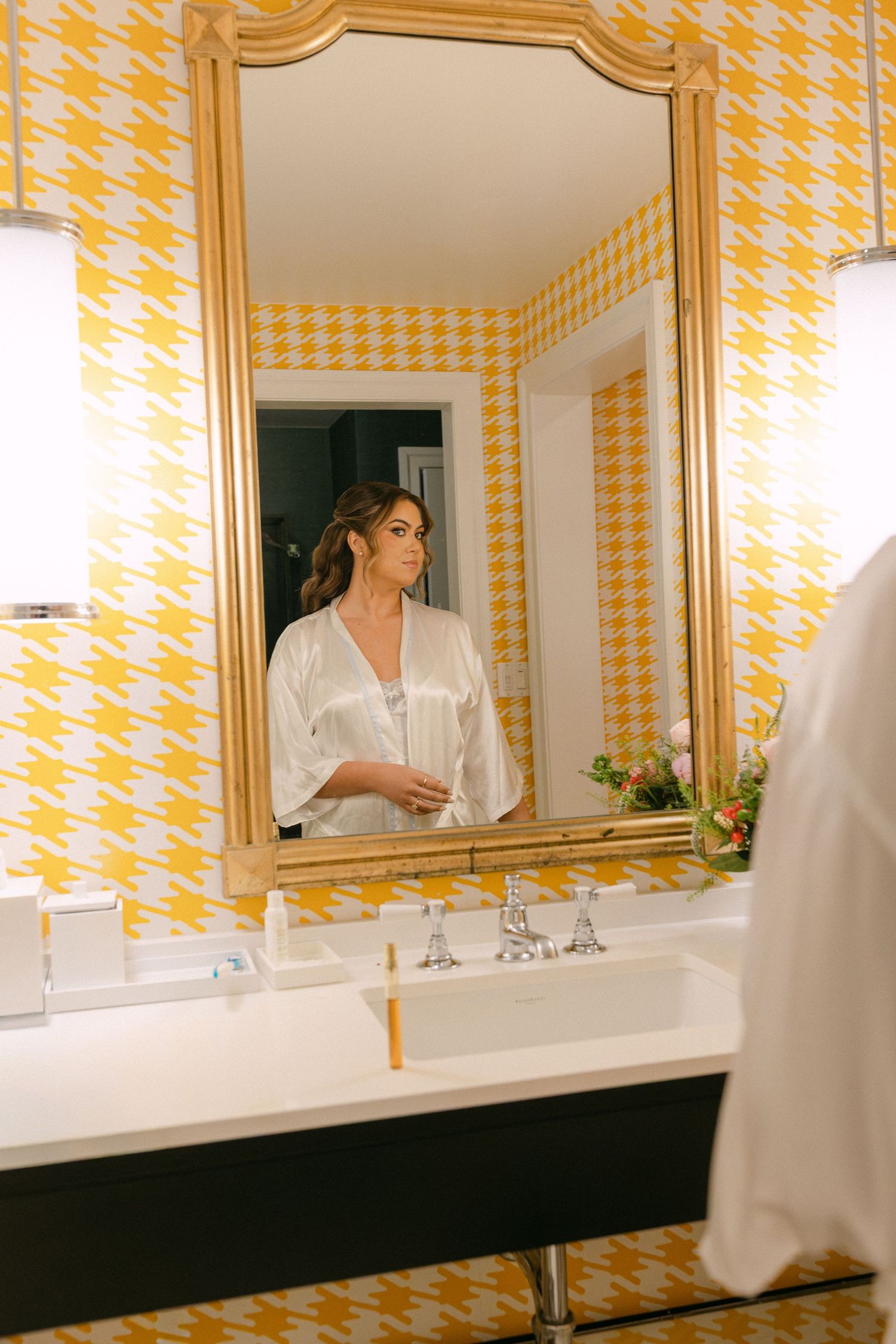 Bride in white satin robe admiring her hair and makeup in a gold-framed mirror with yellow houndstooth wallpaper.