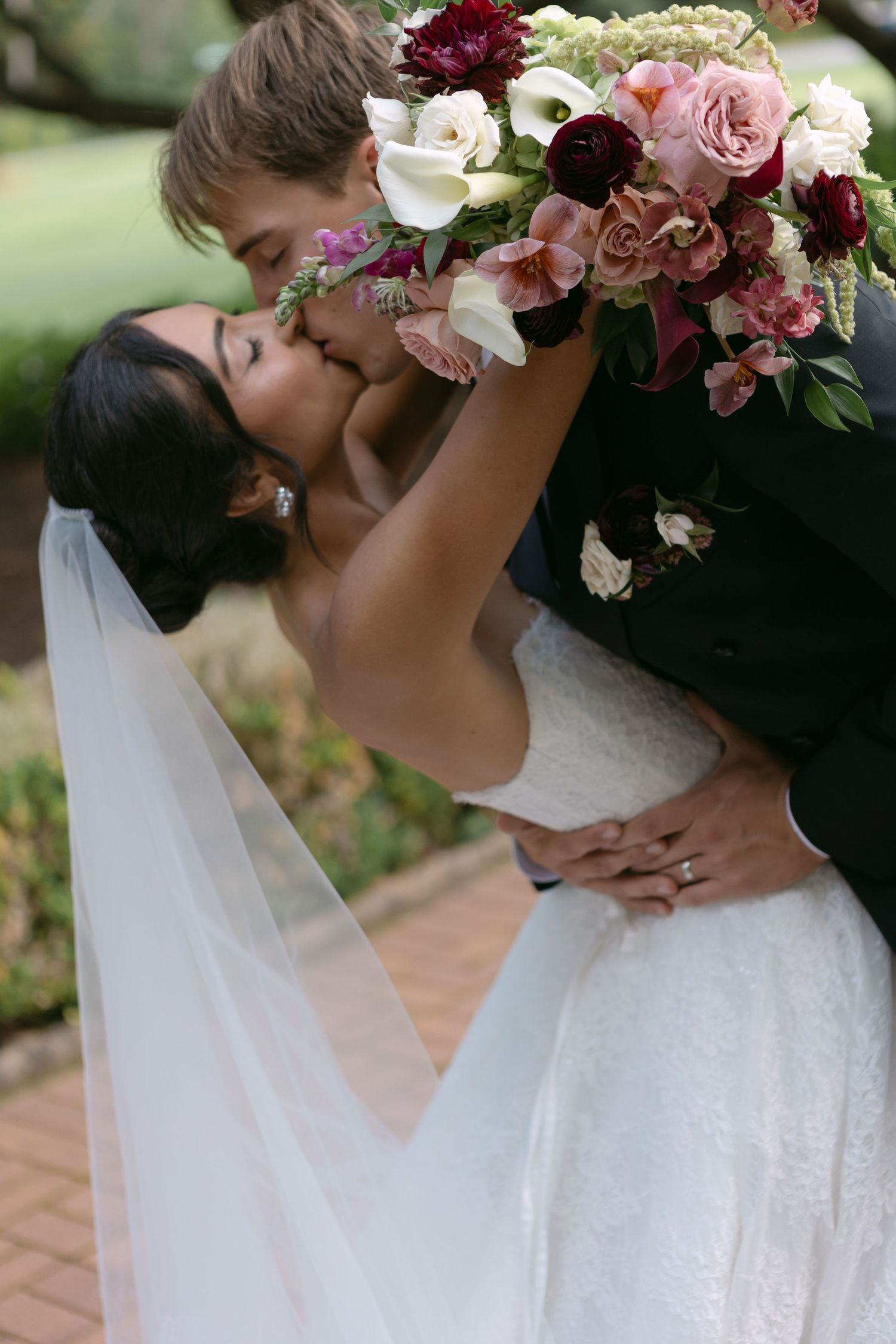 Bride and groom share a romantic kiss while holding a lush bouquet of pink, white, and burgundy flowers outdoors.