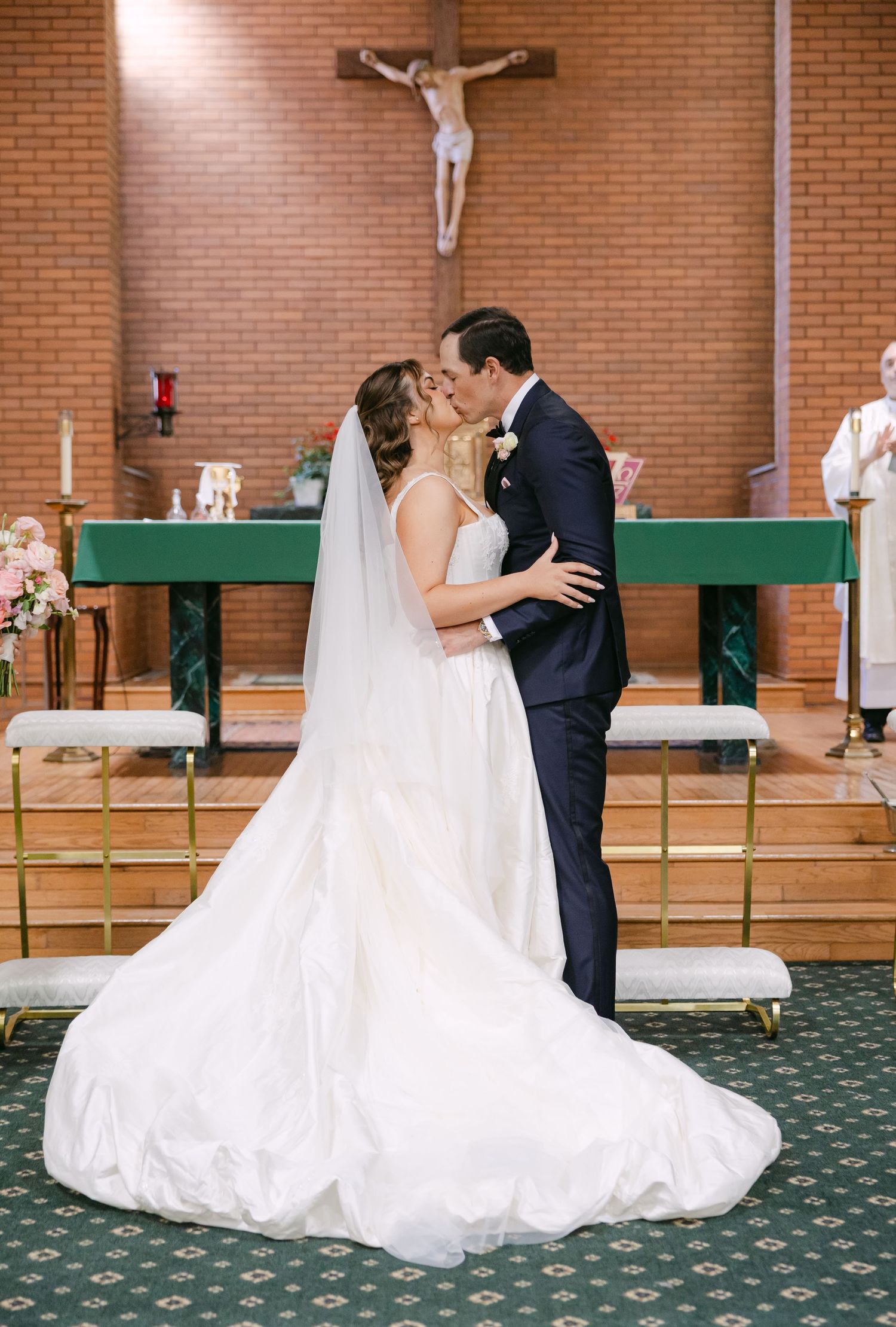 Bride and groom share first kiss at church altar with crucifix backdrop during Catholic wedding ceremony.
