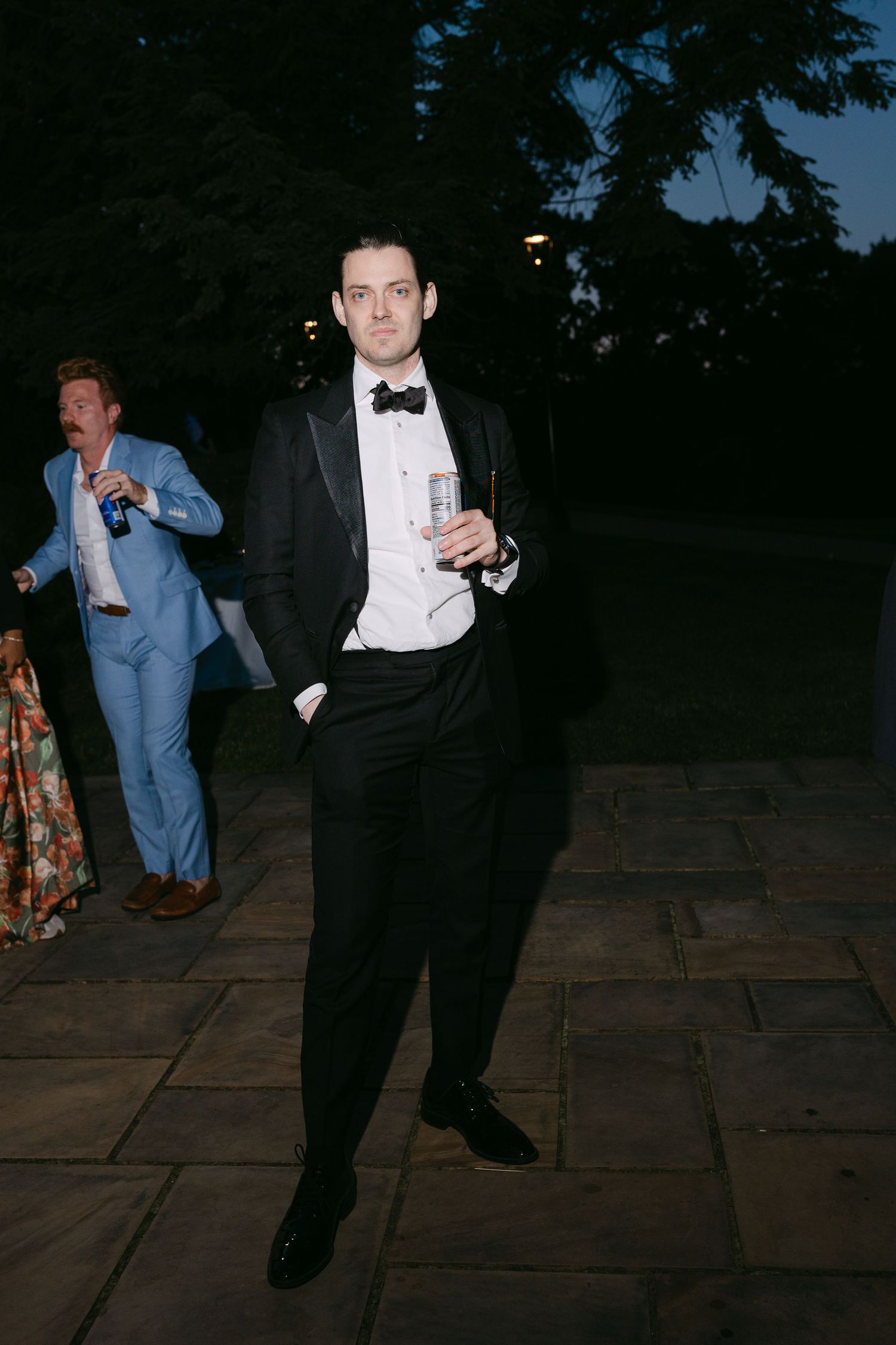 Elegant groom in black tuxedo with bow tie holding a drink outdoors at an evening wedding reception.