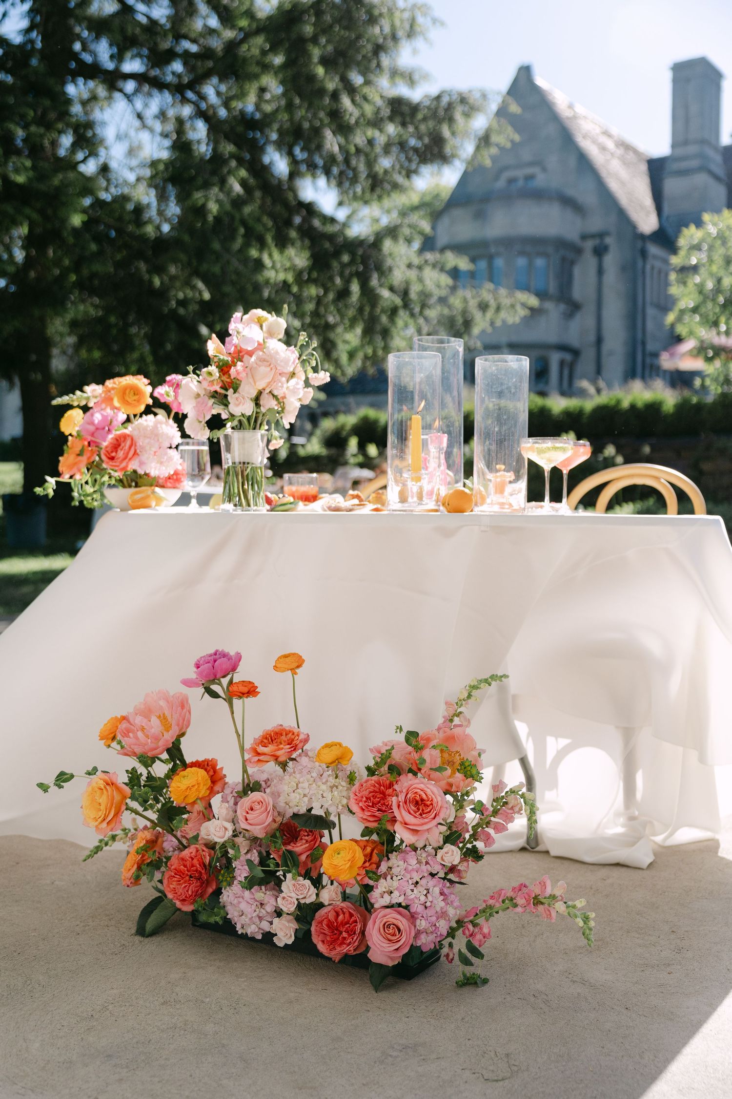 Elegant outdoor wedding sweetheart table with colorful pink and orange floral arrangements, tall glass candles, white tablecloth.