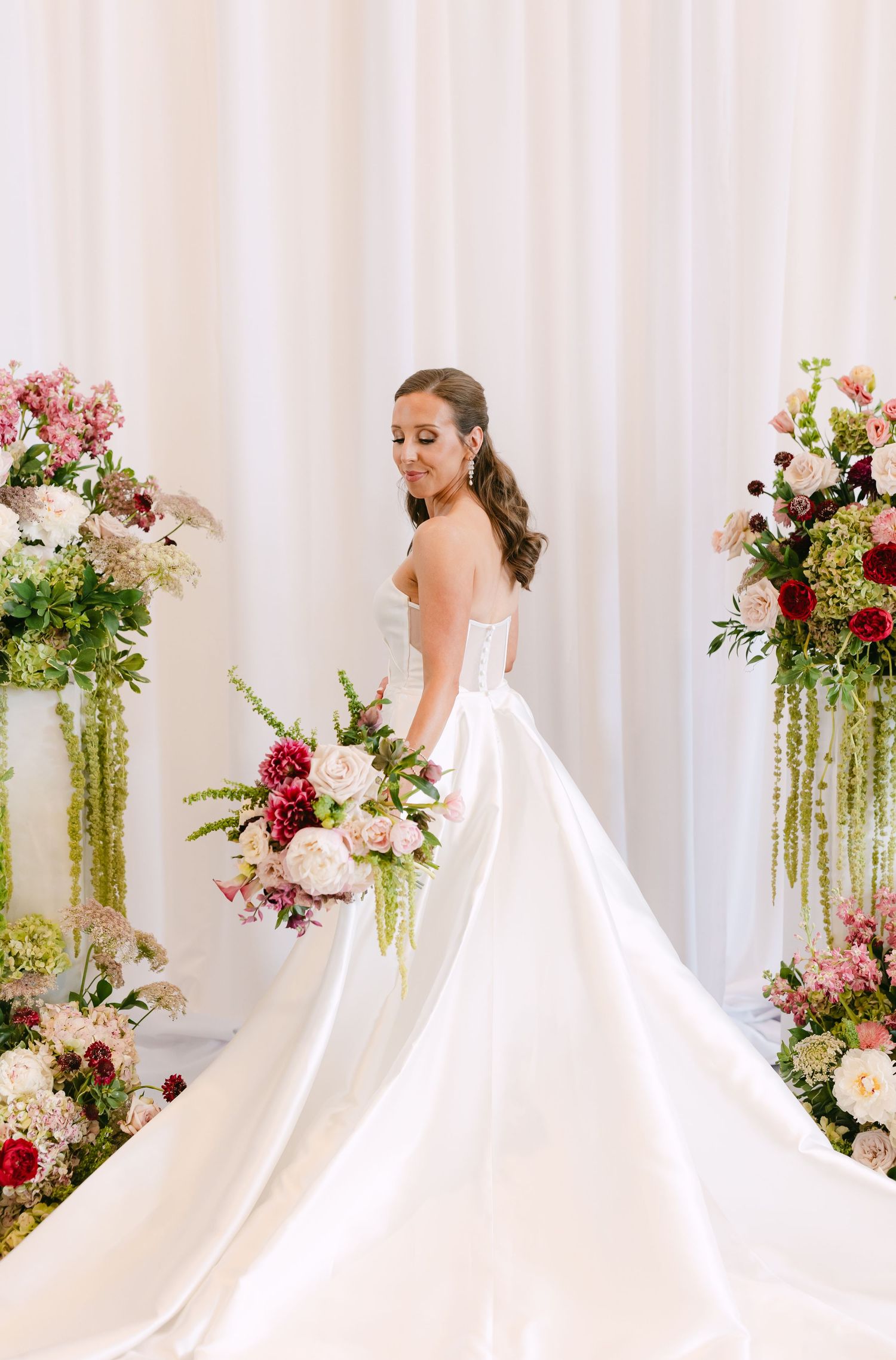 Bride in a strapless satin ballgown holds a lush pink and burgundy bouquet between floral columns.