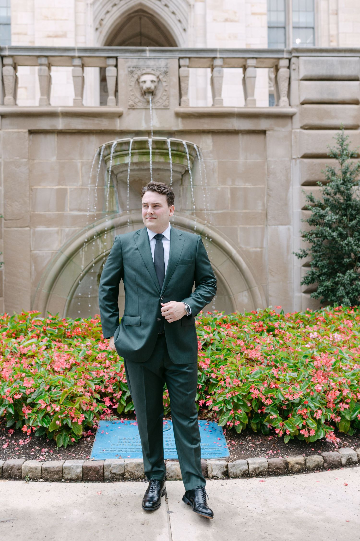 Dapper groom in dark green suit poses before stone fountain surrounded by colorful flowers.