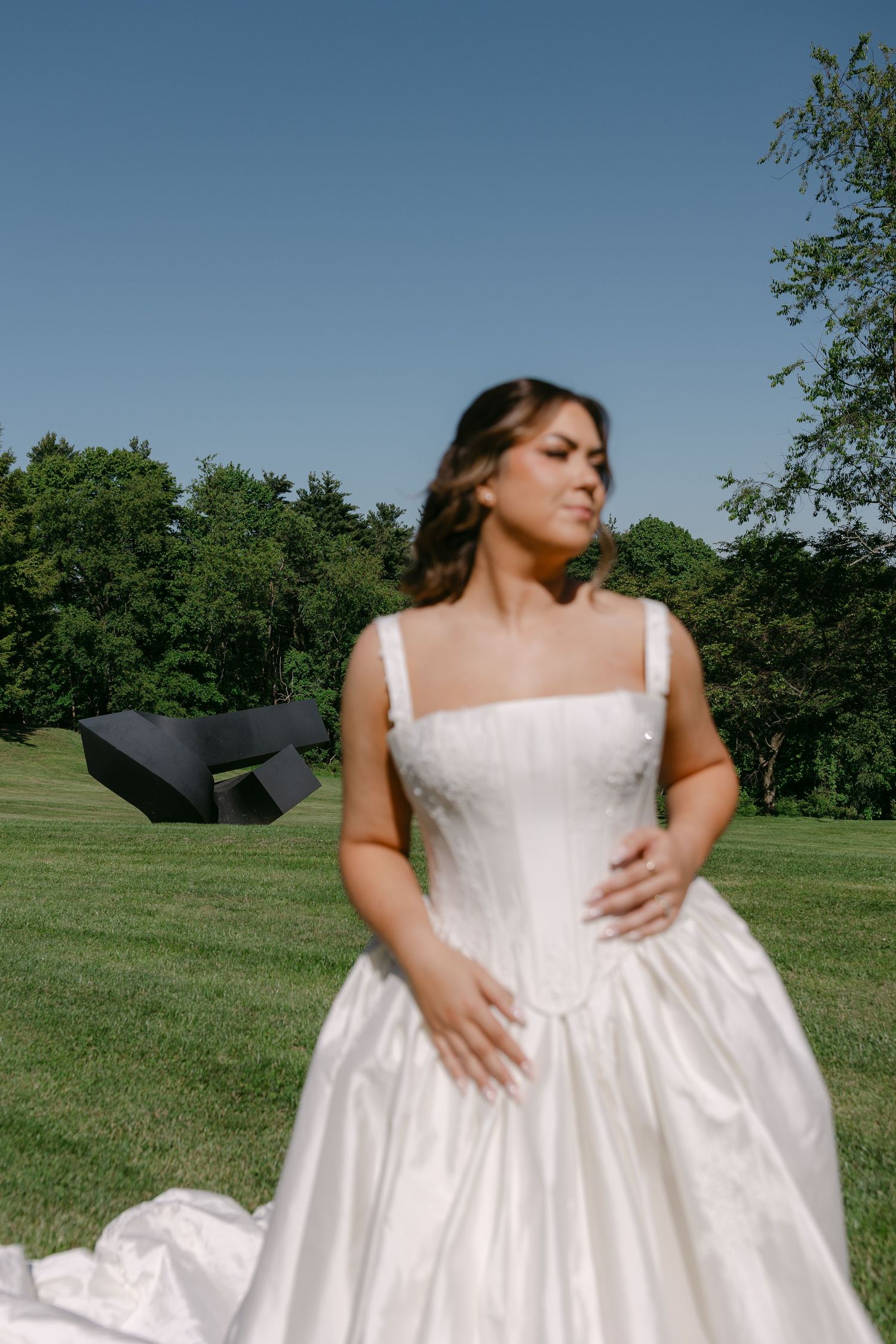 Bride in elegant white ball gown with square neckline stands on lush green lawn outdoors.