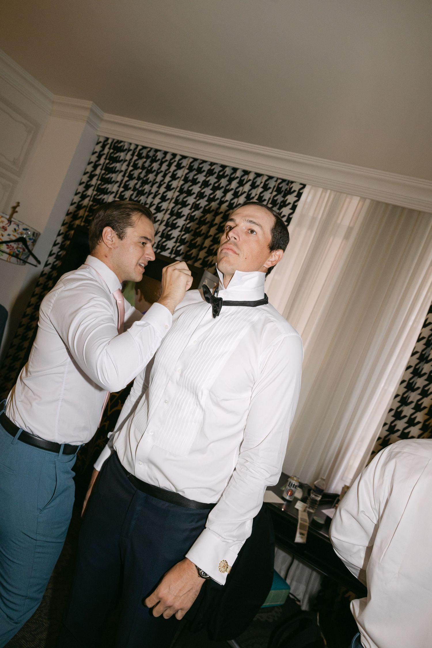 Groomsman helping groom adjust black bow tie while getting dressed in white shirt before wedding.