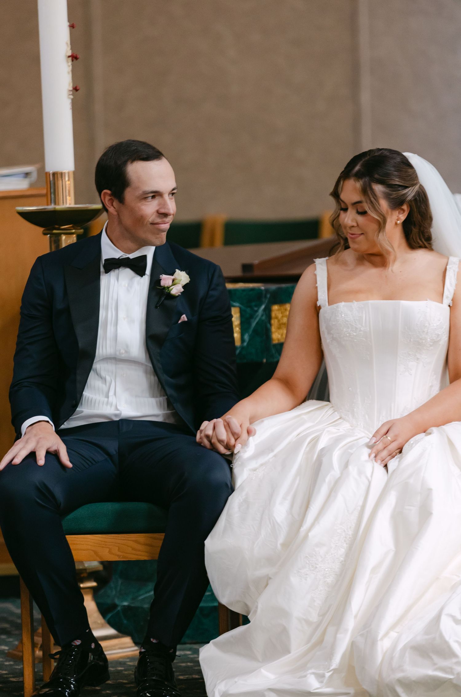 A bride and groom sit together holding hands during their wedding ceremony, dressed in a white gown and navy tuxedo.