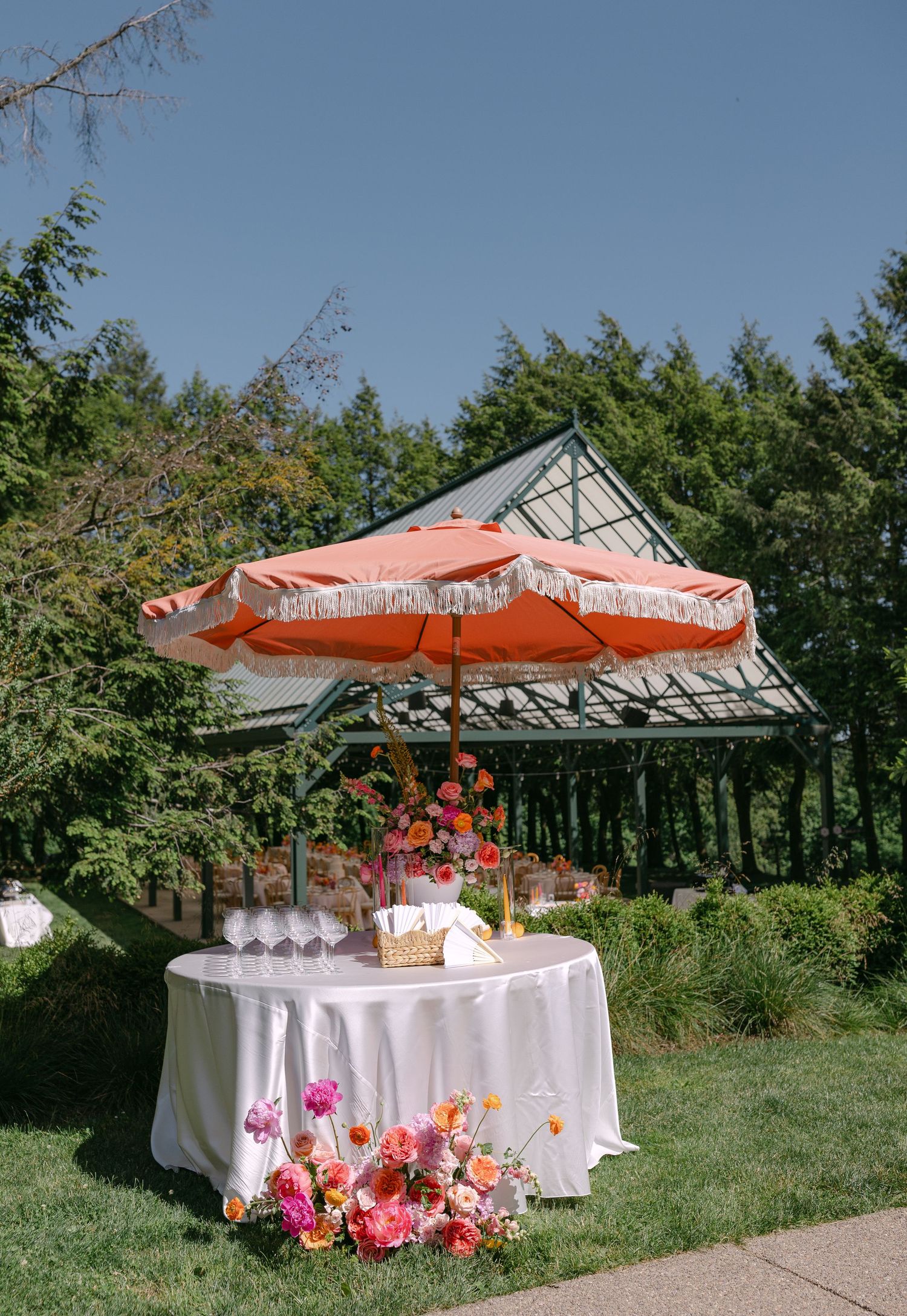 Outdoor wedding cocktail table with orange fringed umbrella and vibrant pink and orange floral arrangements.