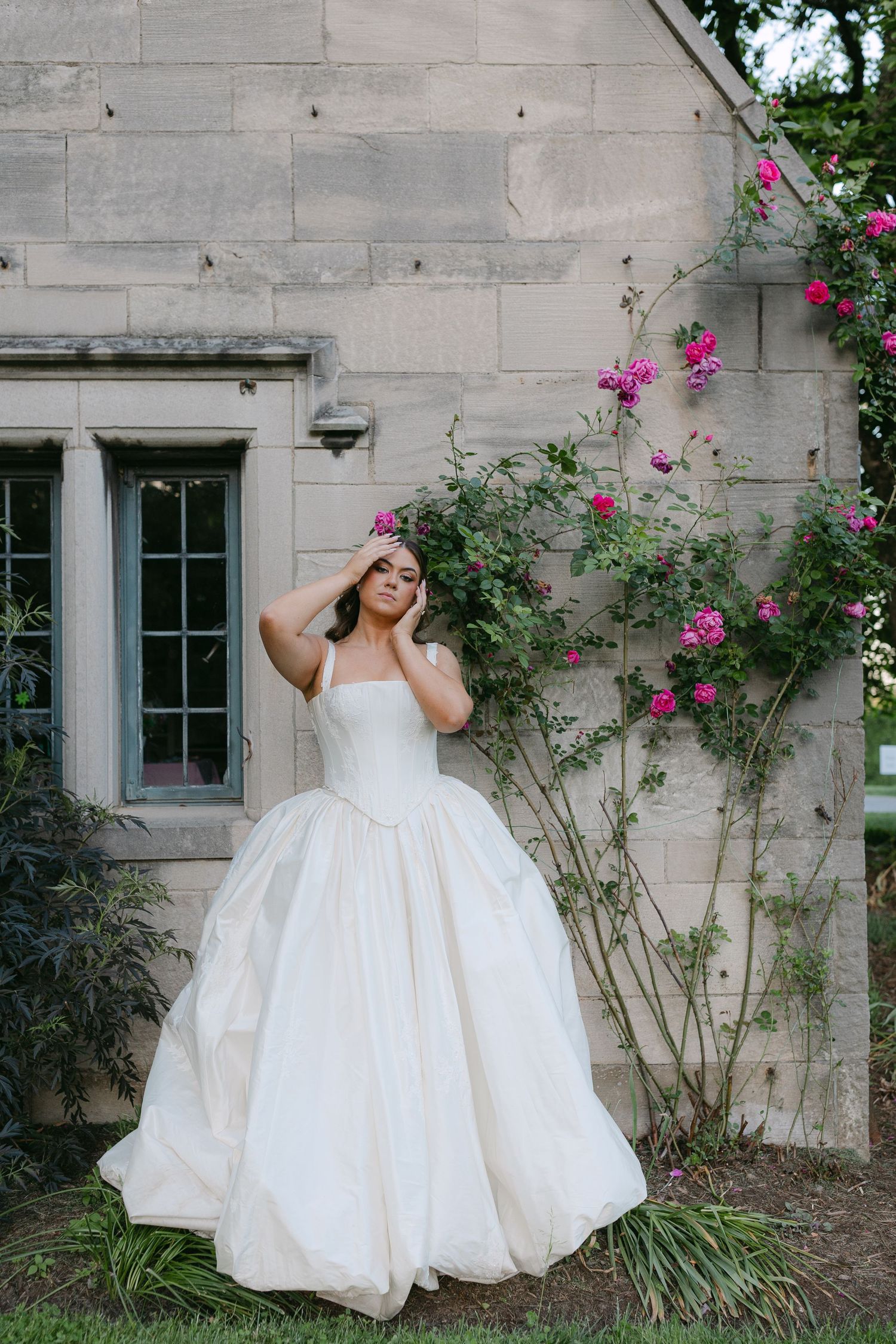 Bride in strapless ivory ball gown poses beside pink climbing roses against stone building.