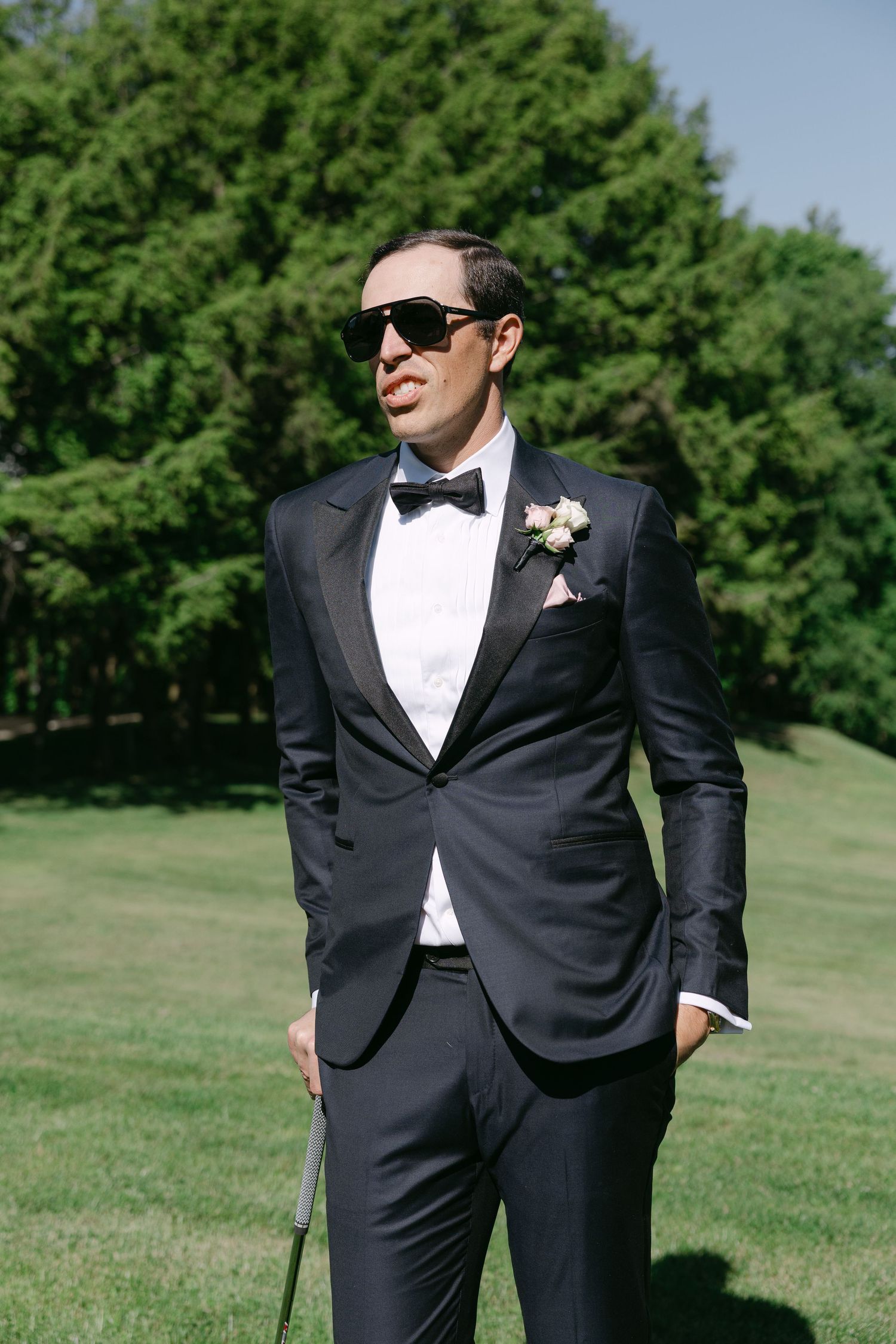 Groom in black tuxedo with bow tie and sunglasses holding a cane outdoors on green lawn.
