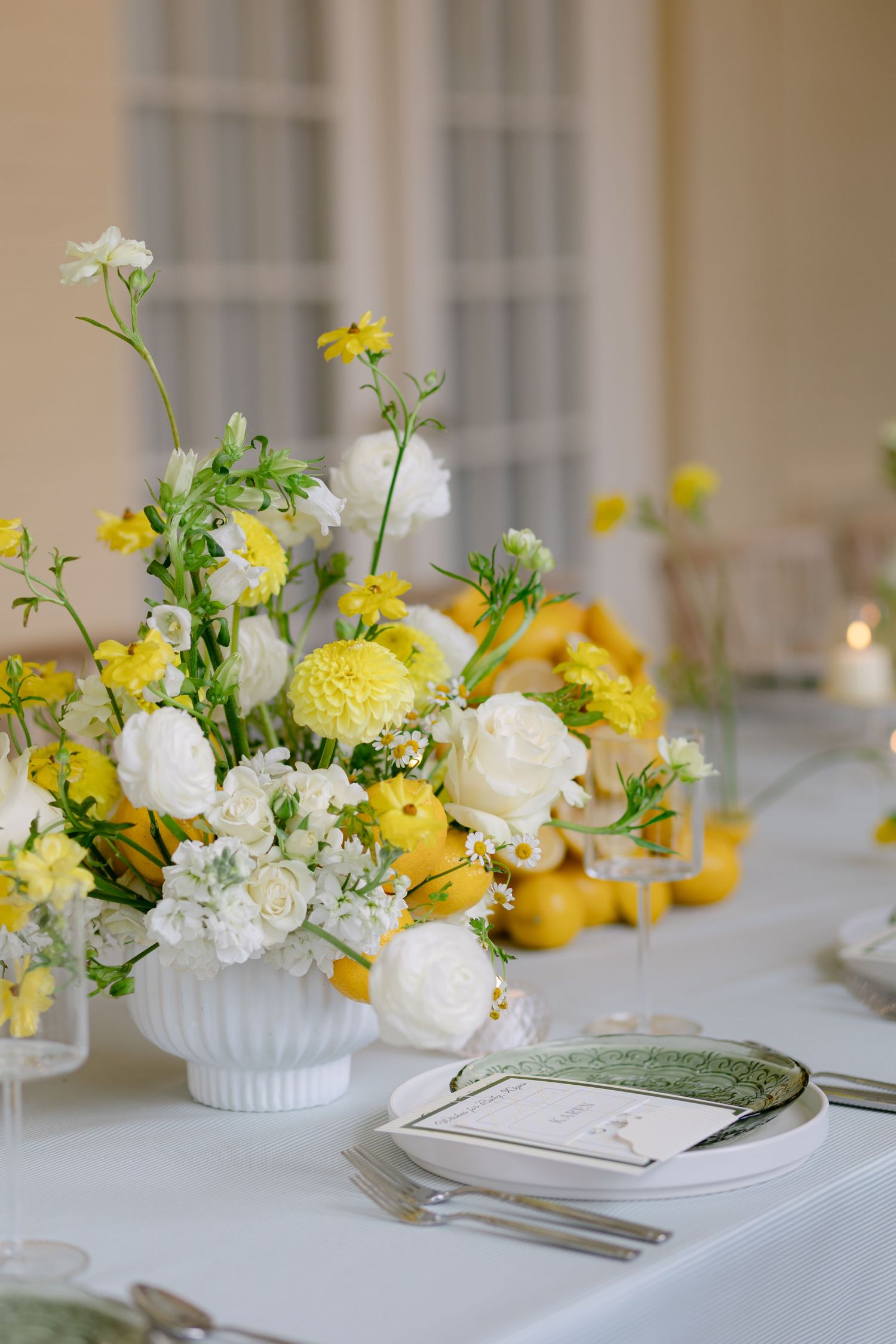 White and yellow floral wedding centerpiece with lemons in a white pedestal bowl on a reception table.