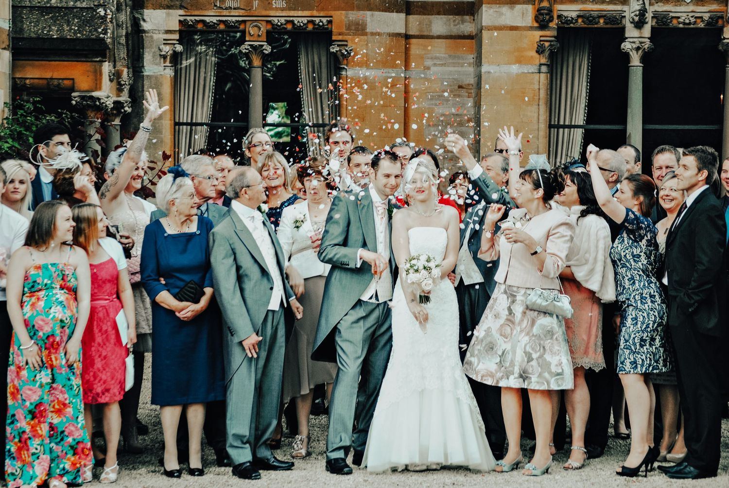 A large wedding party gathers for a group photo outside a historic building with ornate architecture.