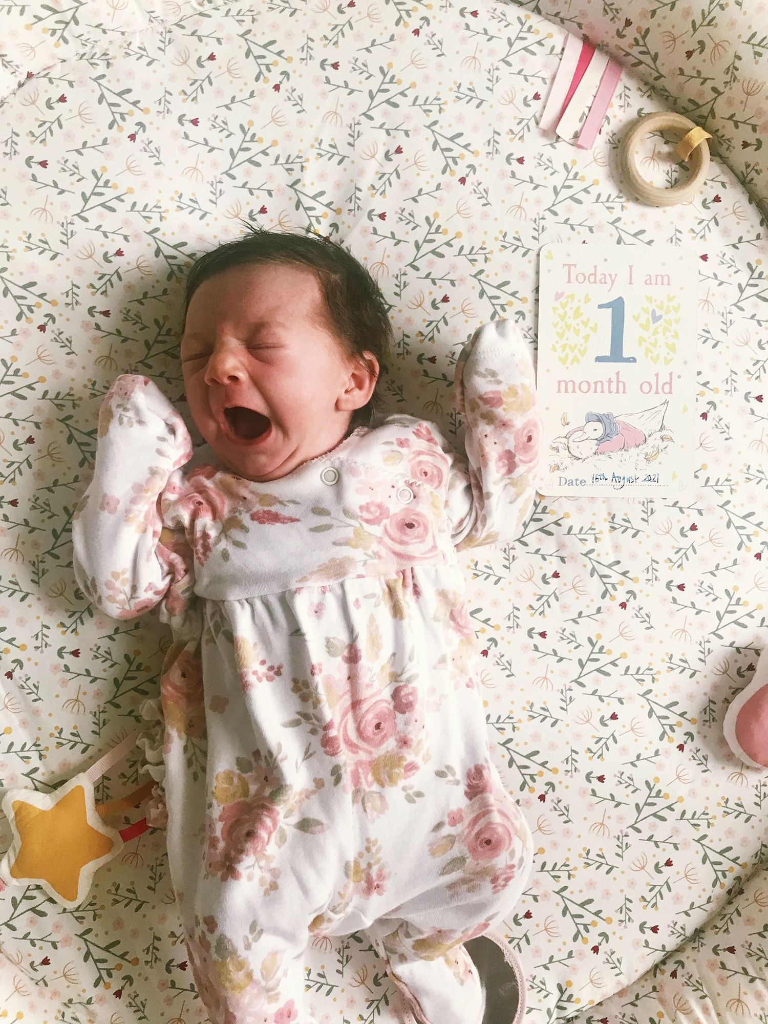 One month old baby in floral dress crying on a patterned blanket with milestone card.