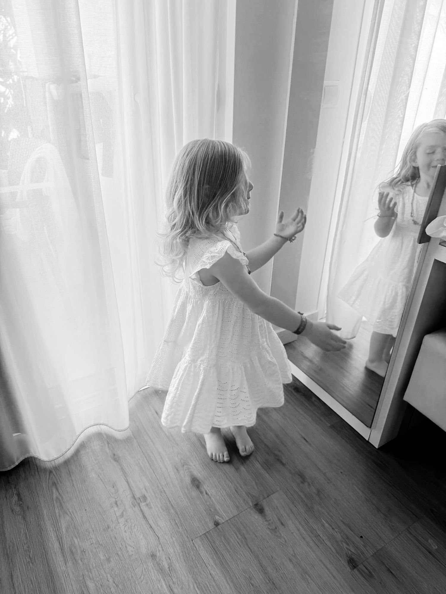 Child in white dress plays near curtains and mirror in sunlit room.
